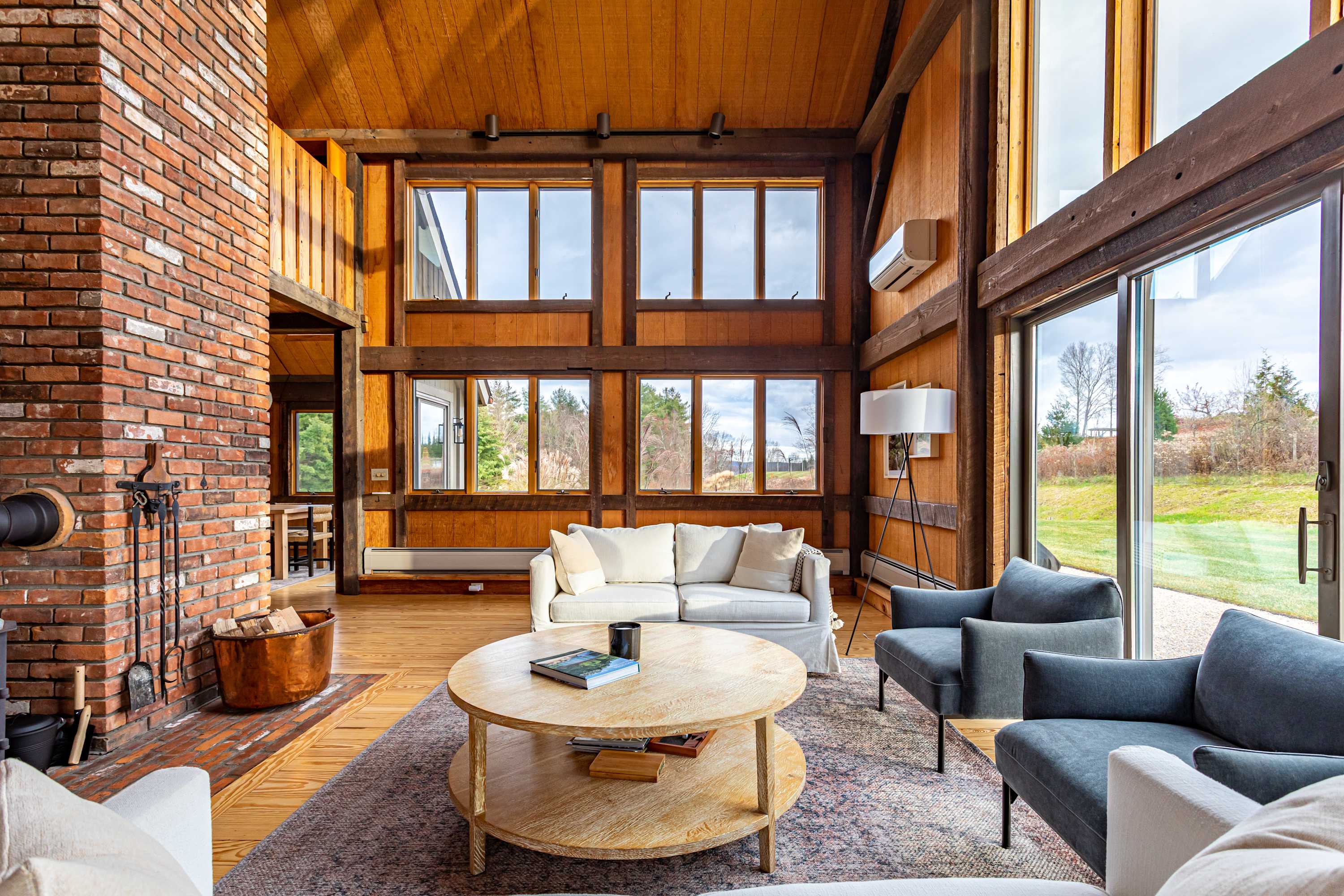 Grand living room with a towering brick fireplace, exposed wooden beams, and high ceilings, featuring comfortable white sofas and gray armchairs on a patterned rug, with large windows providing ample natural light.