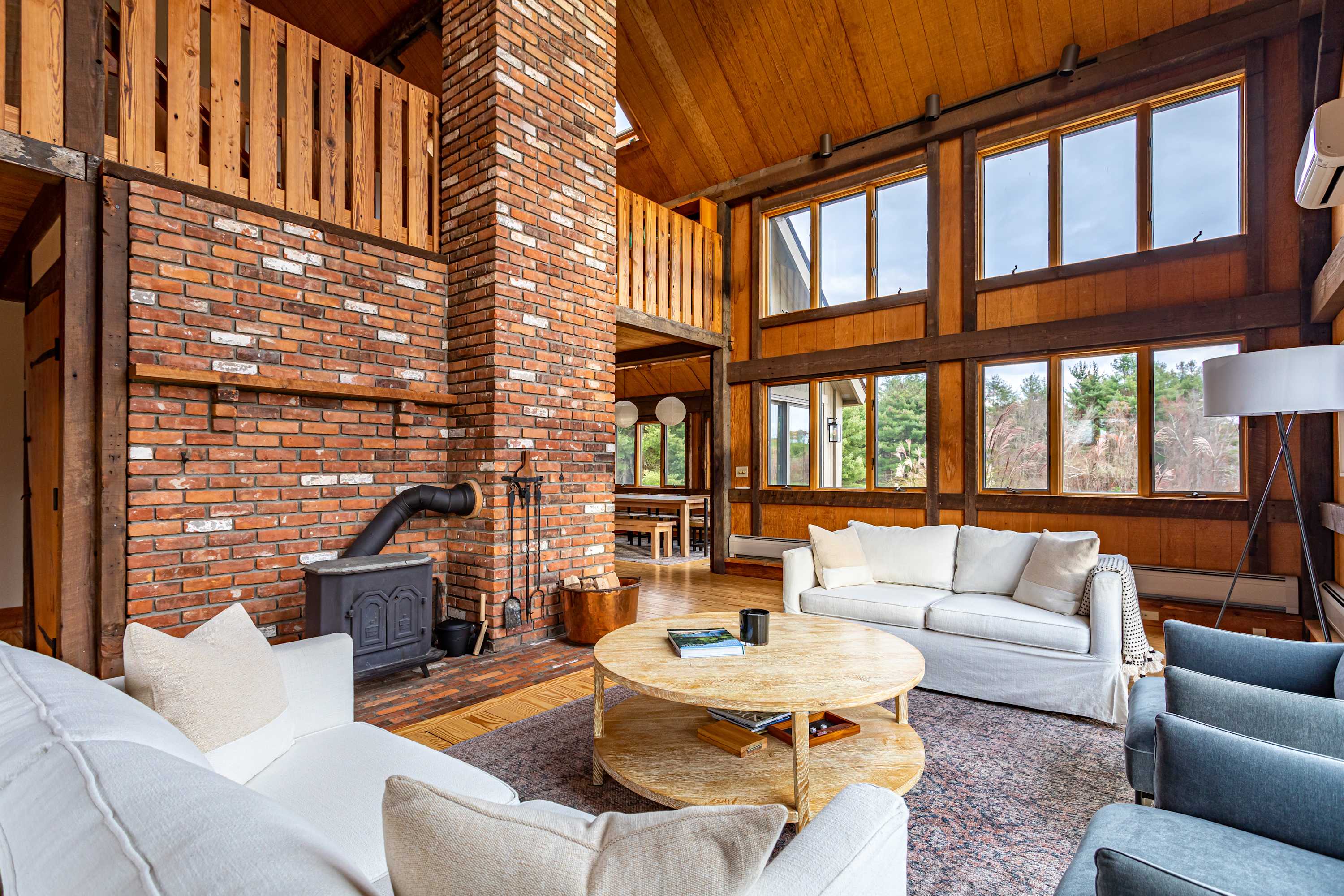 A spacious living room with a white sofa and two gray armchairs centered around a round wooden coffee table on a patterned rug. A large brick fireplace with a wood-burning stove anchors one wall, while expansive windows offer views of the surrounding greenery. The room boasts high wood-paneled ceilings and exposed wooden beams, with a lofted area visible above.