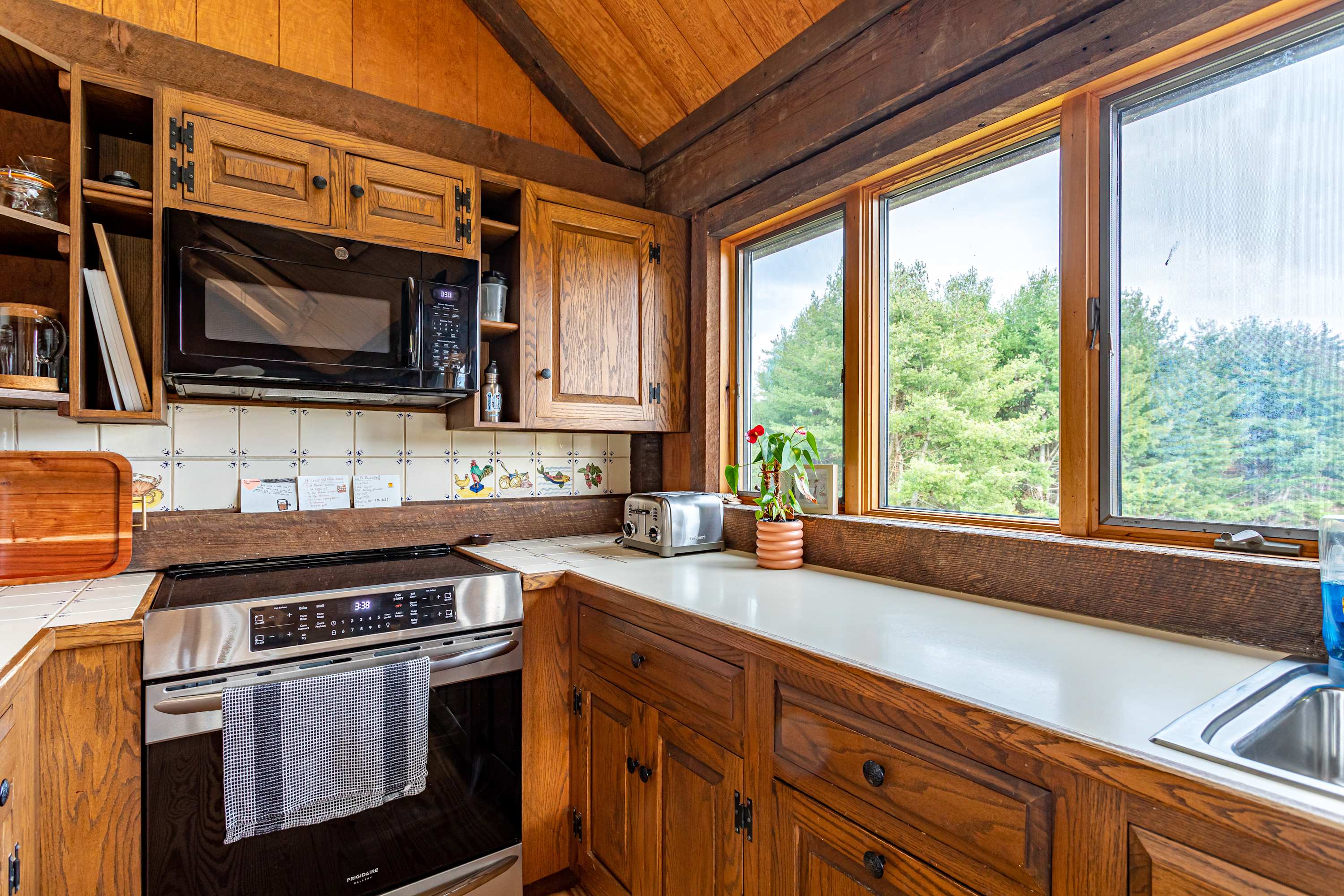 A close-up view of a rustic kitchen showing wooden cabinetry, light countertops, and a stainless steel electric range with a microwave above it. A large window provides natural light and a view of trees, while the backsplash features decorative tiles. The room has a wood-paneled vaulted ceiling.