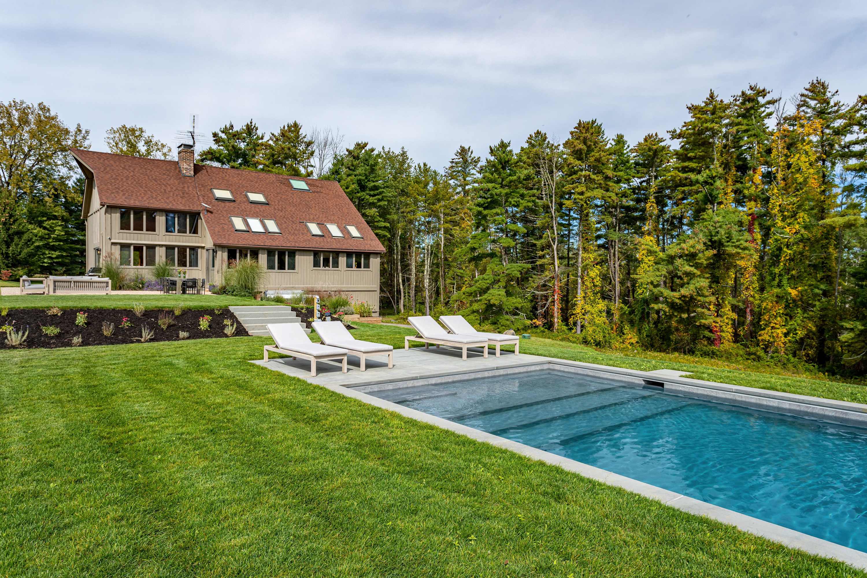 Rectangular swimming pool with lounge chairs on a manicured green lawn, with the multi-level house visible in the background, surrounded by a mix of evergreen and deciduous trees showing autumn colors.