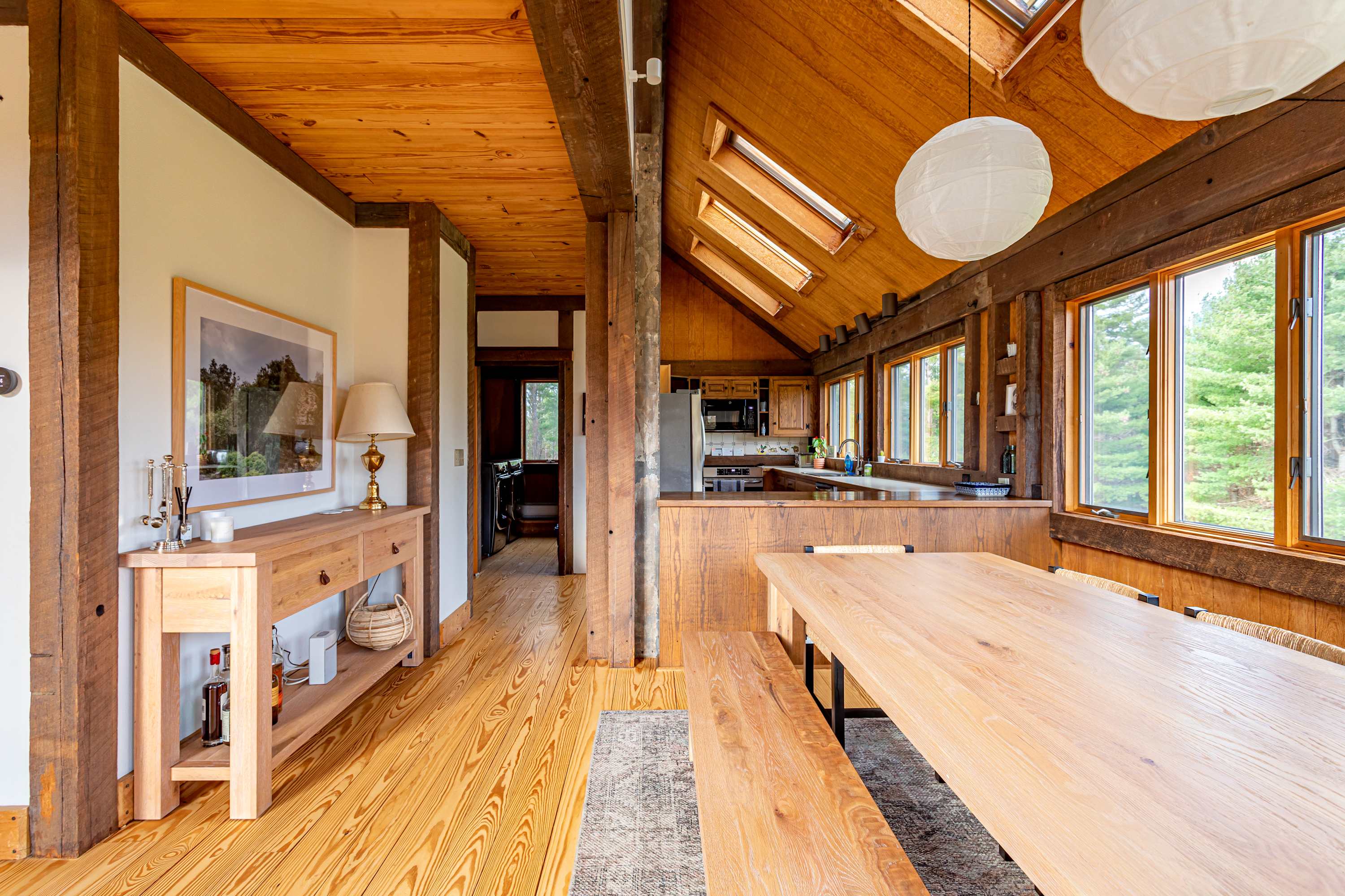 A rustic dining area with a long wooden table and benches, complemented by a patterned rug. The space features warm wood-paneled walls and ceilings with exposed beams, and large windows offering views of the outdoors. A wooden console table with a lamp and framed art is visible to the left, and the kitchen is partially visible in the background.