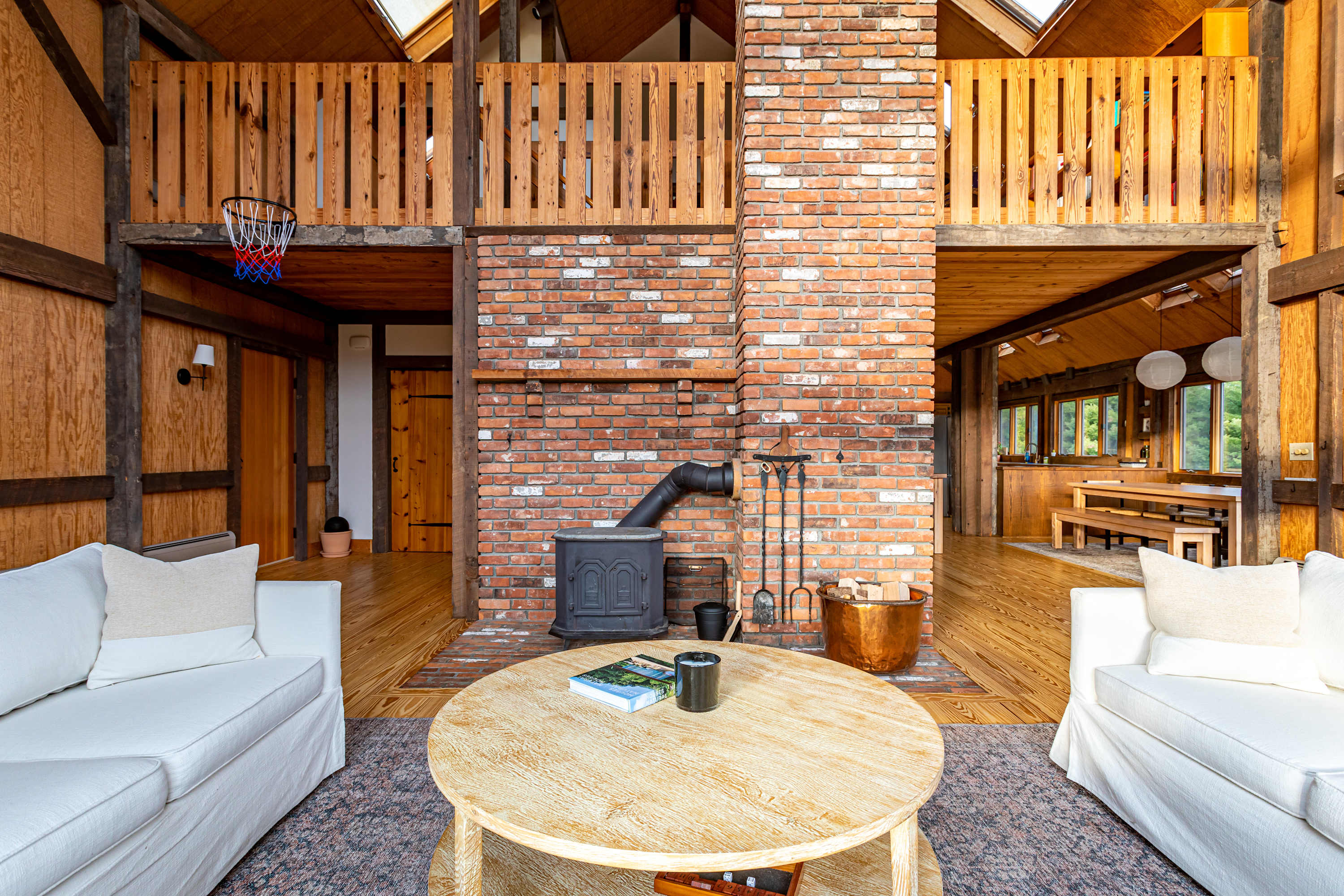 A cozy living area features two white sofas facing each other across a round wooden coffee table, set on a patterned rug. A prominent brick fireplace with a wood-burning stove serves as a central focal point, with a mini basketball hoop visible on the upper level. The room showcases rustic wood-paneled walls and ceilings with exposed beams.