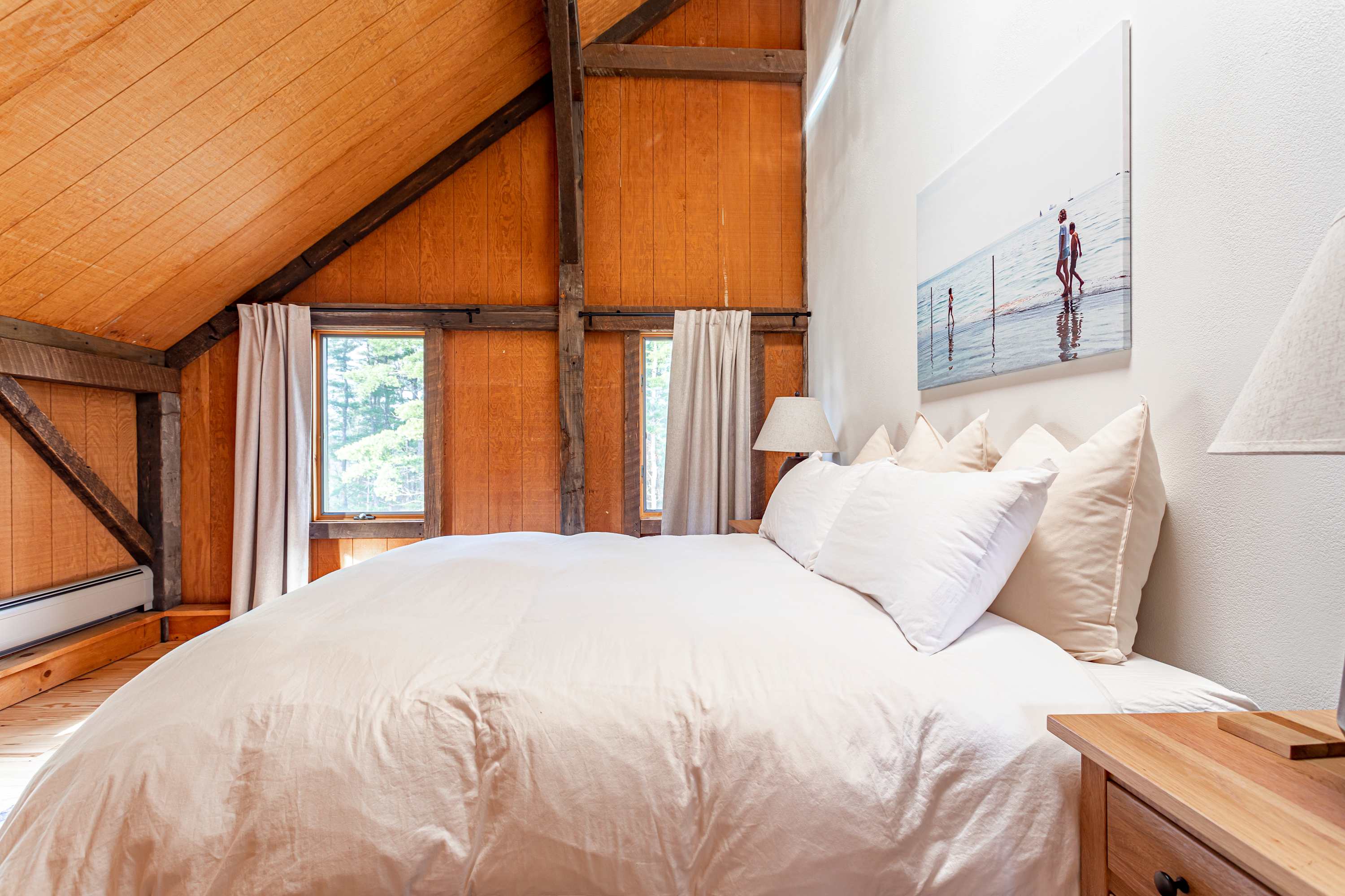 Bedroom with a bed featuring cream-colored bedding, set against a white wall with a large artwork. The room has sloped wooden ceilings and exposed beams, with two windows on the side.