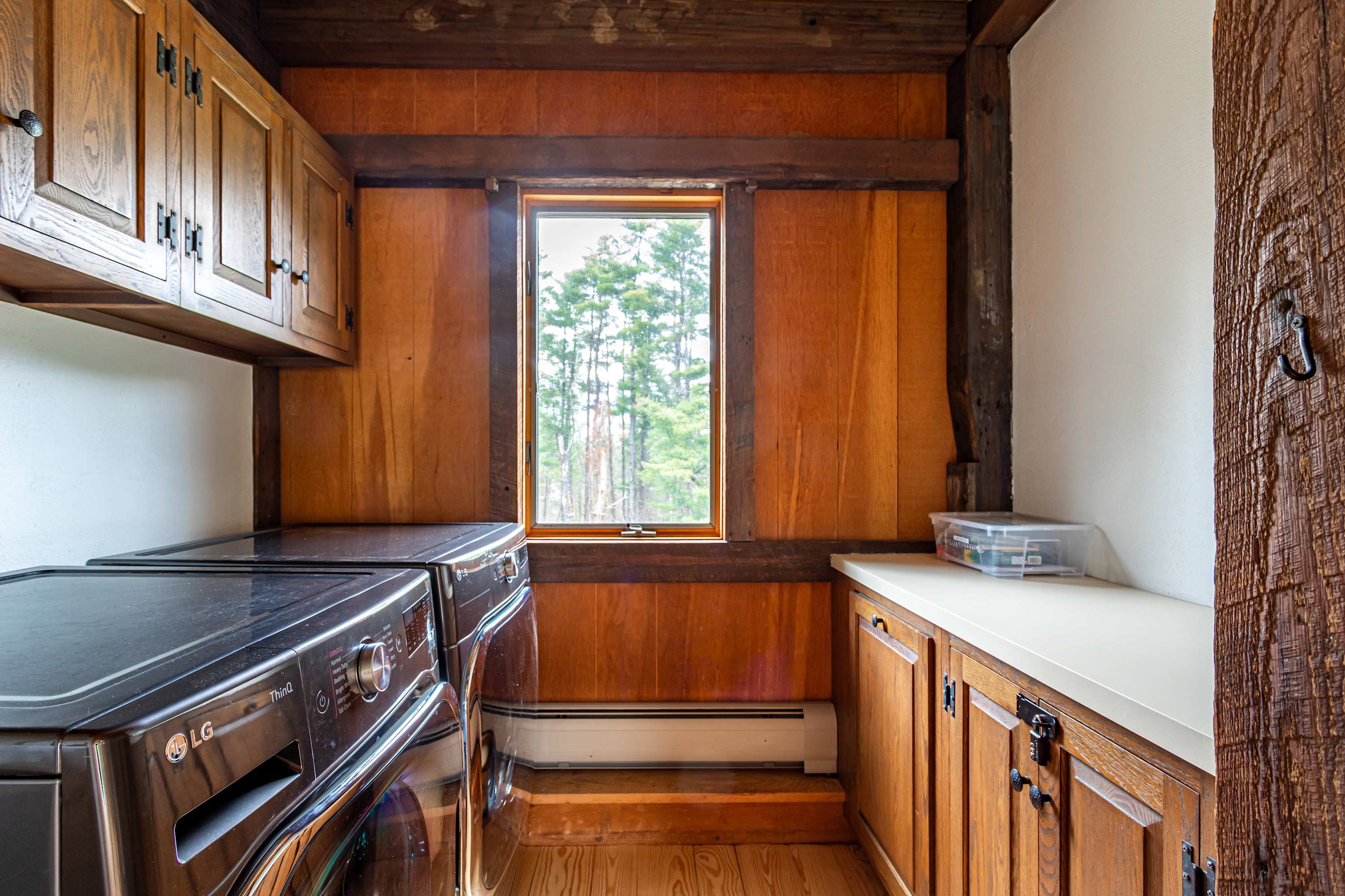 Rustic laundry room with wood-paneled walls and cabinets, featuring a dark LG washer and dryer set, a small window overlooking trees, and a white countertop with storage below.