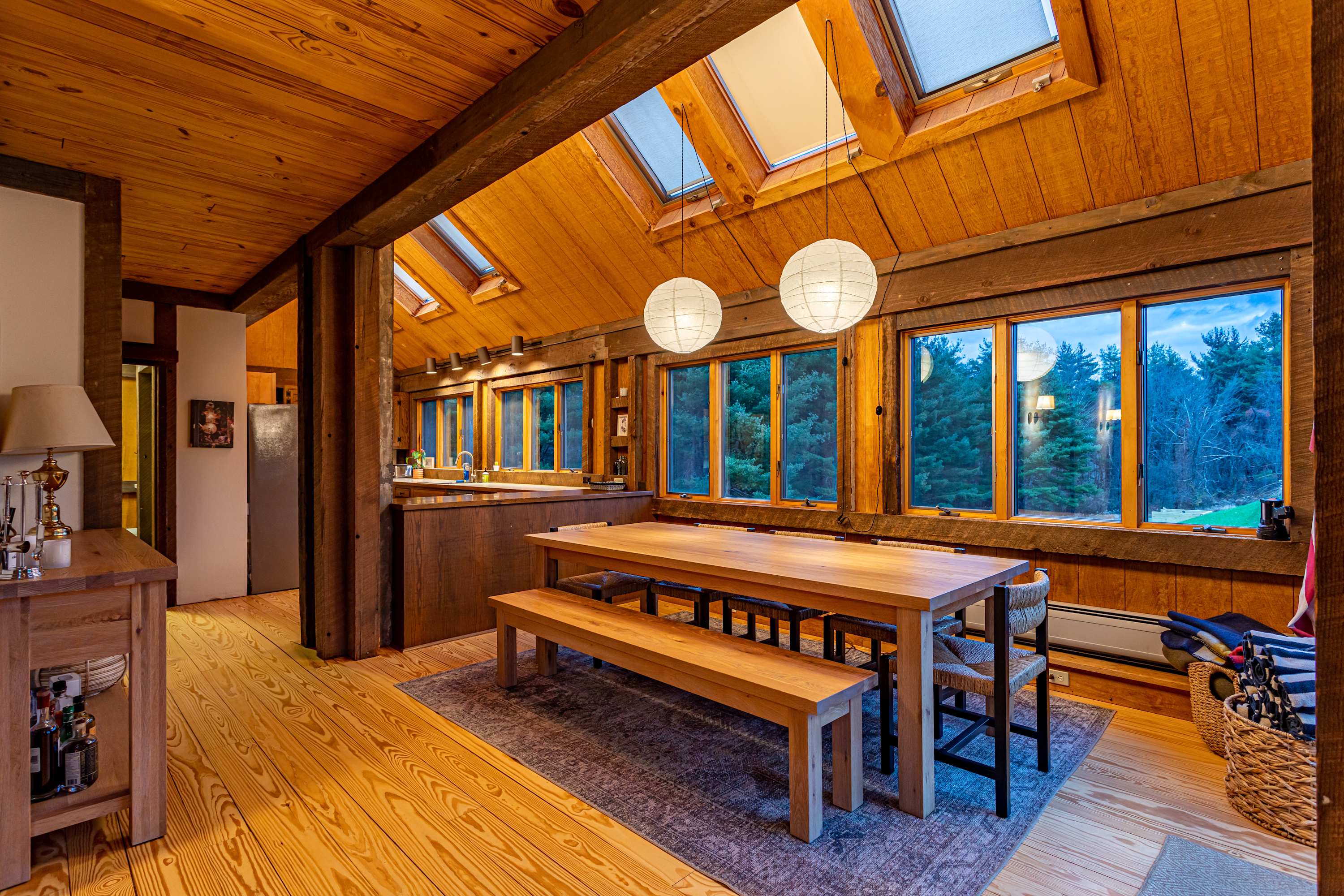 Bright dining area with a long wooden table and benches, illuminated by two modern pendant lights and multiple skylights, adjacent to a kitchen counter. The room features extensive wood paneling and large windows overlooking trees.