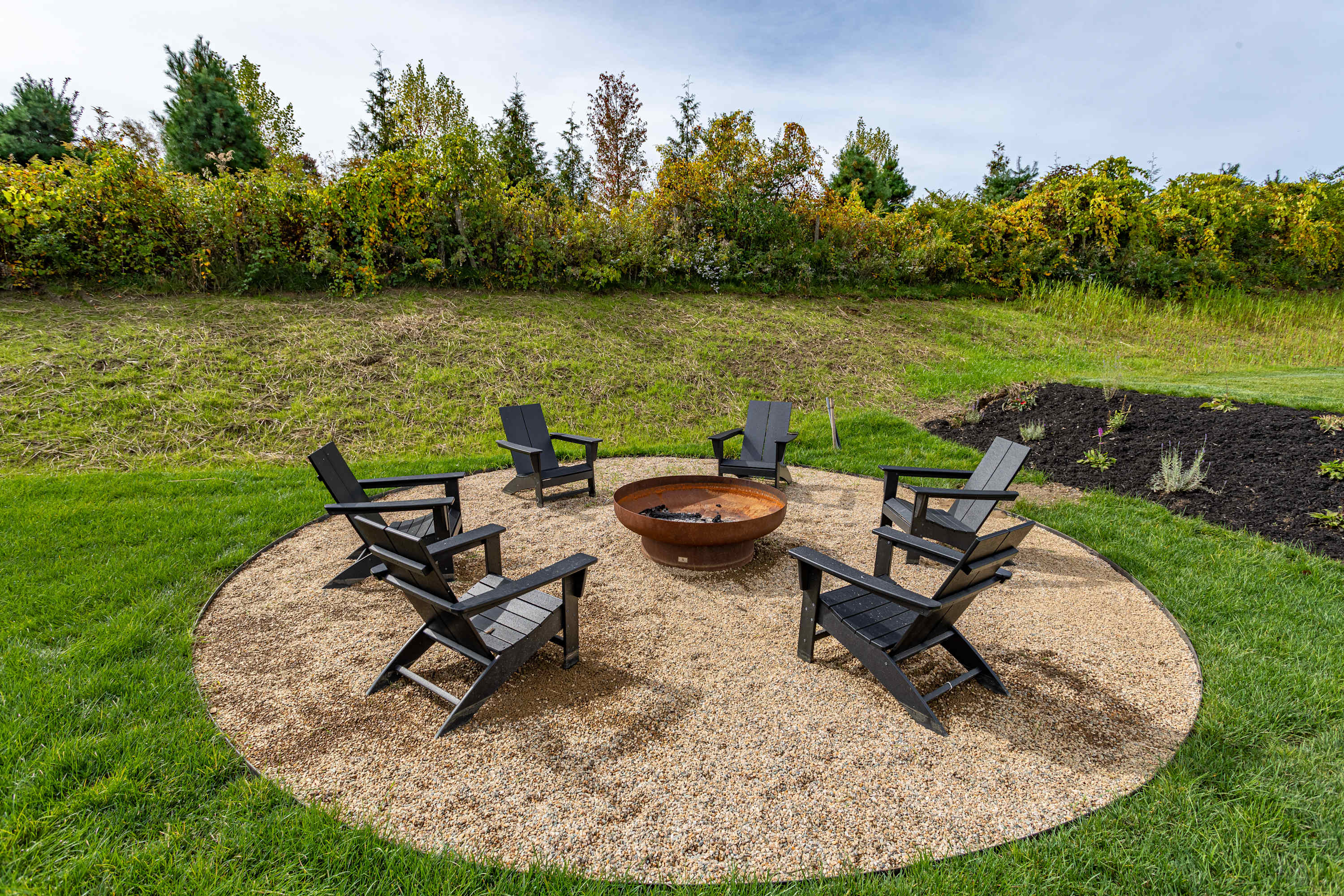 Circular gravel area with a large metal fire pit at its center, surrounded by six black Adirondack chairs, all set on a green lawn with a sloped, treed background.