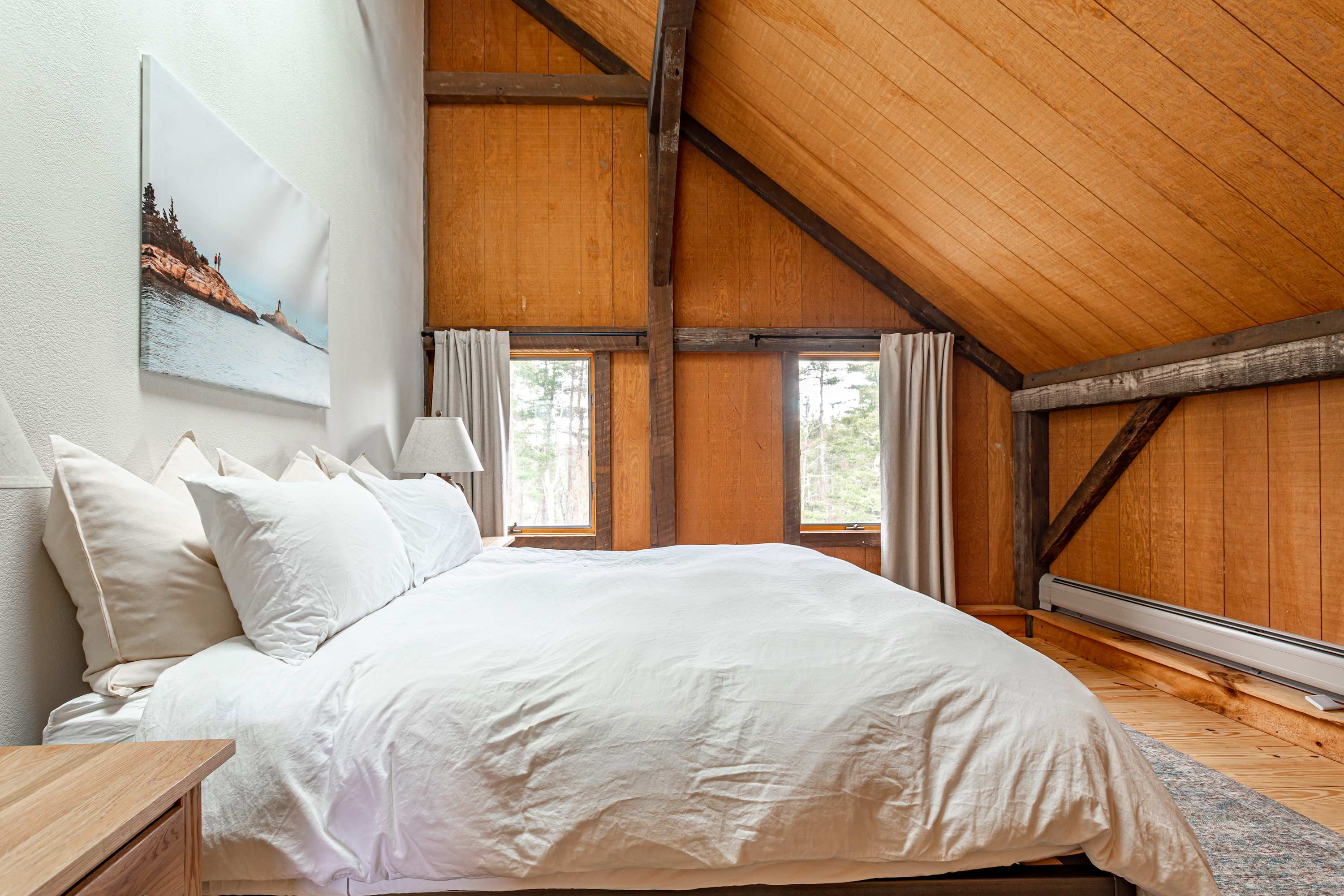 Cozy bedroom with a white-duvet bed, sloped wooden ceilings, and exposed beams. Two small windows with light curtains offer glimpses of the outdoors.