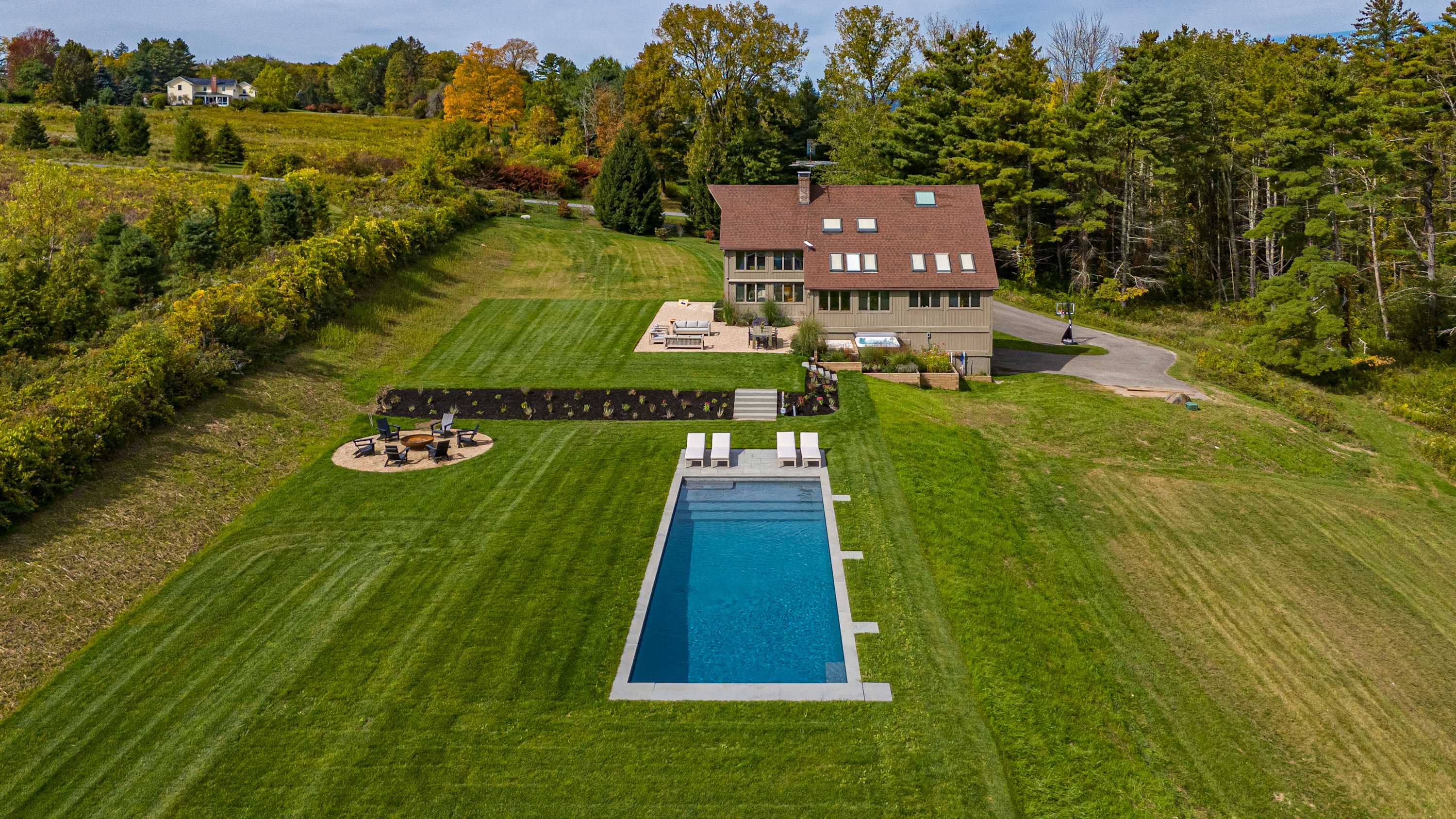 Aerial view of a large house with a brown roof and multiple skylights, situated on a sprawling green lawn with a rectangular swimming pool, a fire pit area, and surrounded by lush trees and rolling hills.