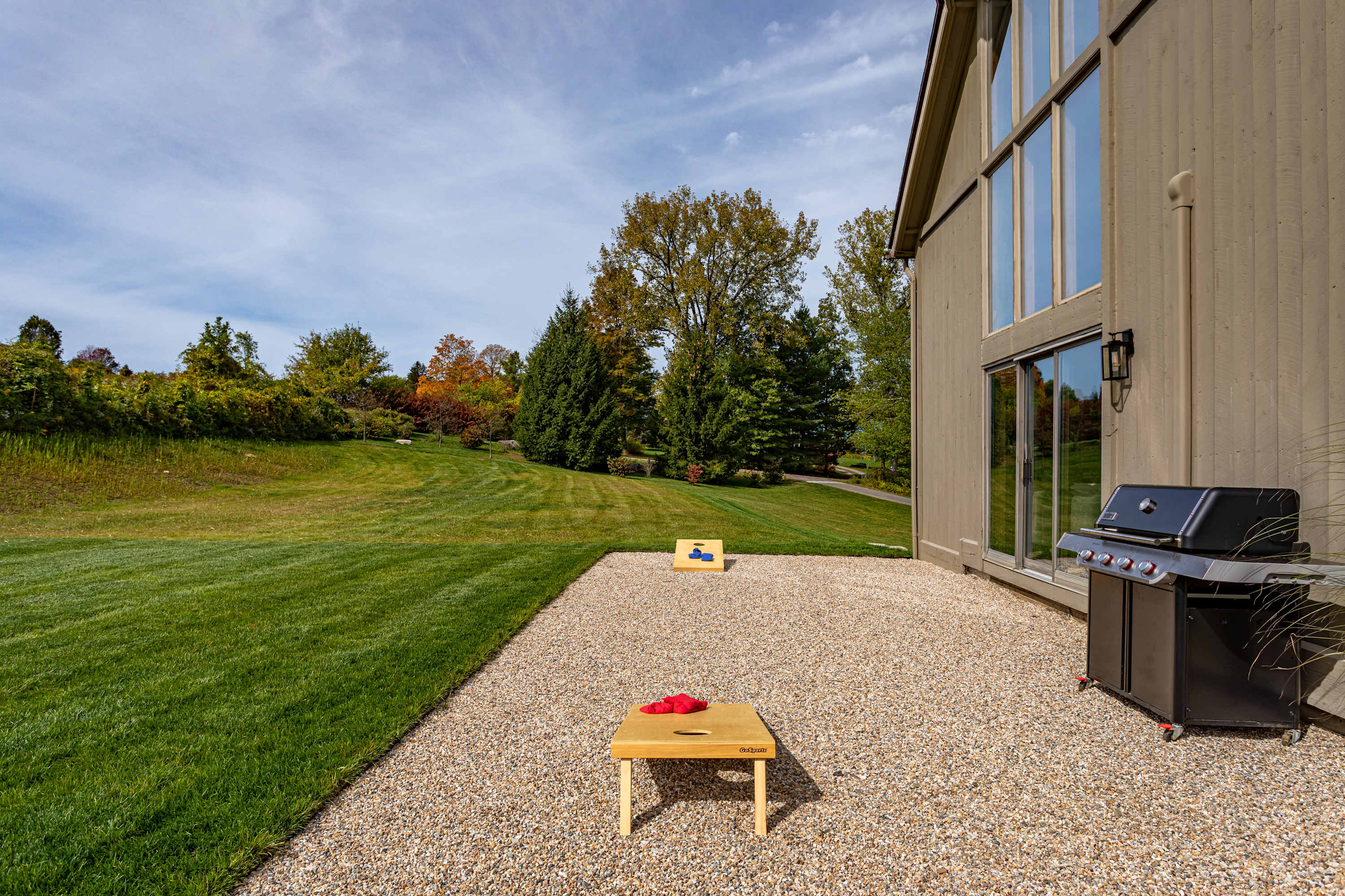 Outdoor patio area with light gravel, featuring a black BBQ grill, a cornhole game setup, and a view of the house's large windows and a grassy hillside.