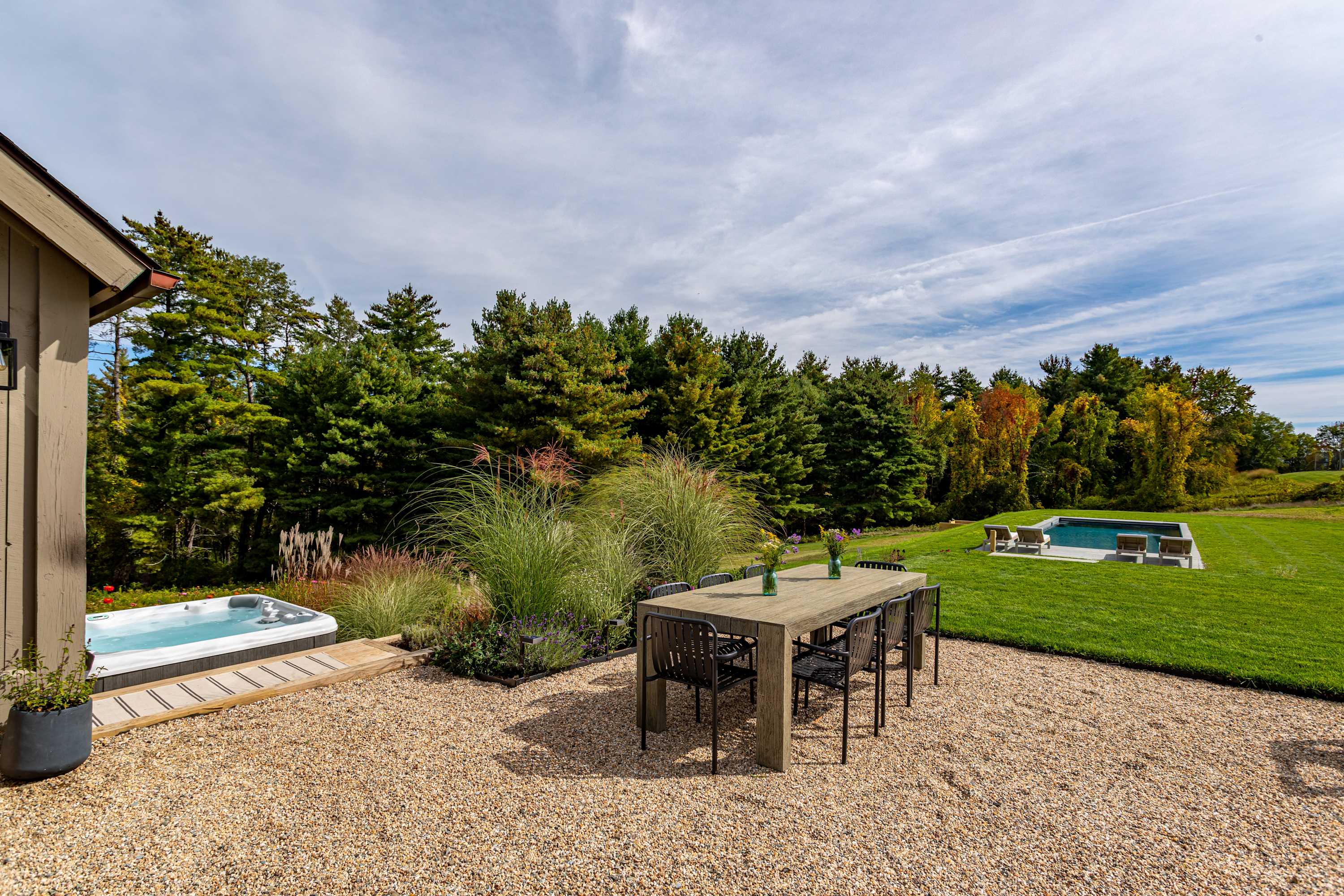 Spacious gravel patio featuring a large dark outdoor dining table with chairs, a hot tub nestled in a wooden surround, and landscaped plants, with the swimming pool visible in the distance.