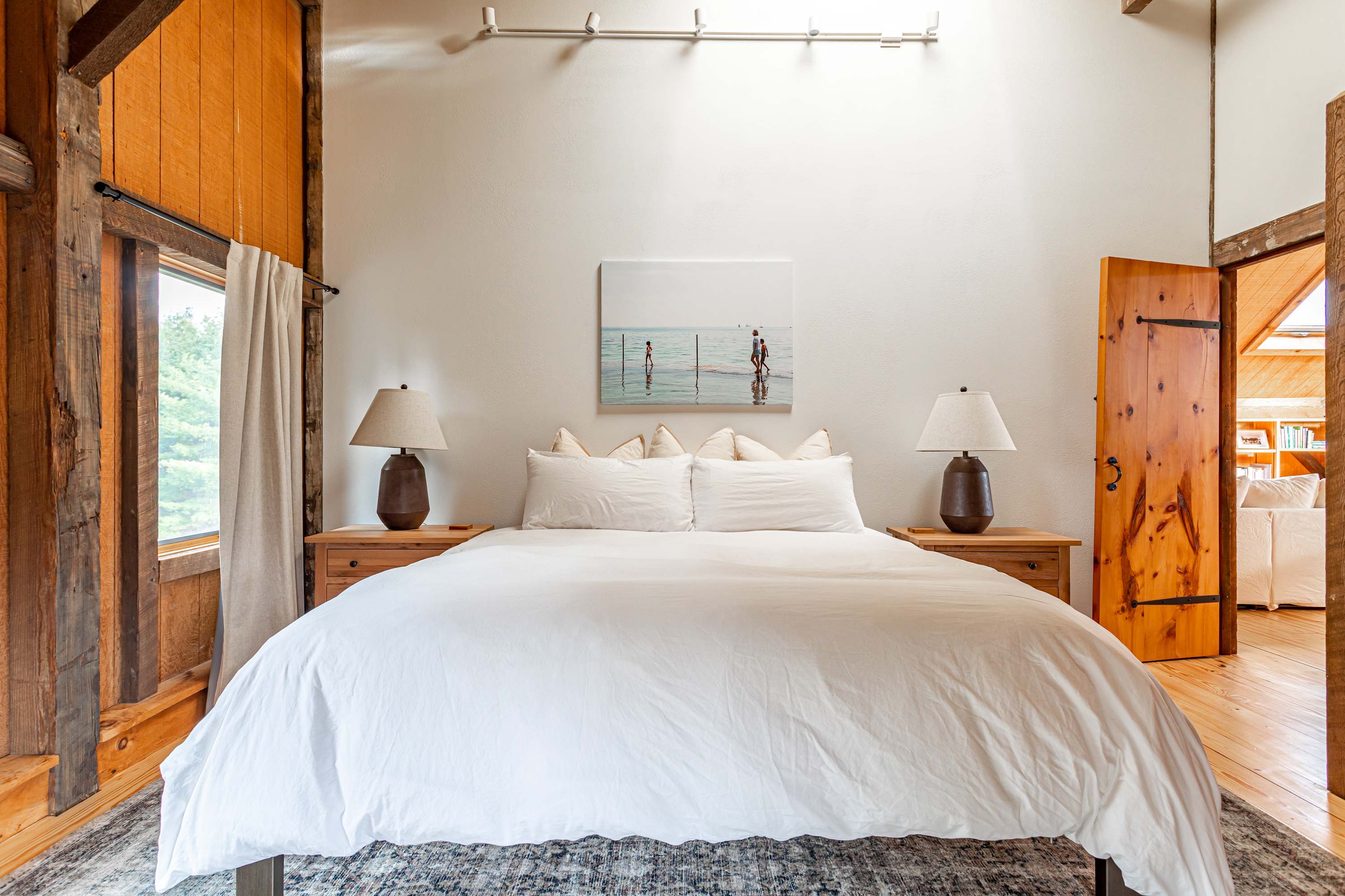 Spacious bedroom featuring a king-sized bed with white bedding, flanked by wooden nightstands with dark ceramic lamps. A large artwork hangs above the headboard, complementing the rustic wooden beams.