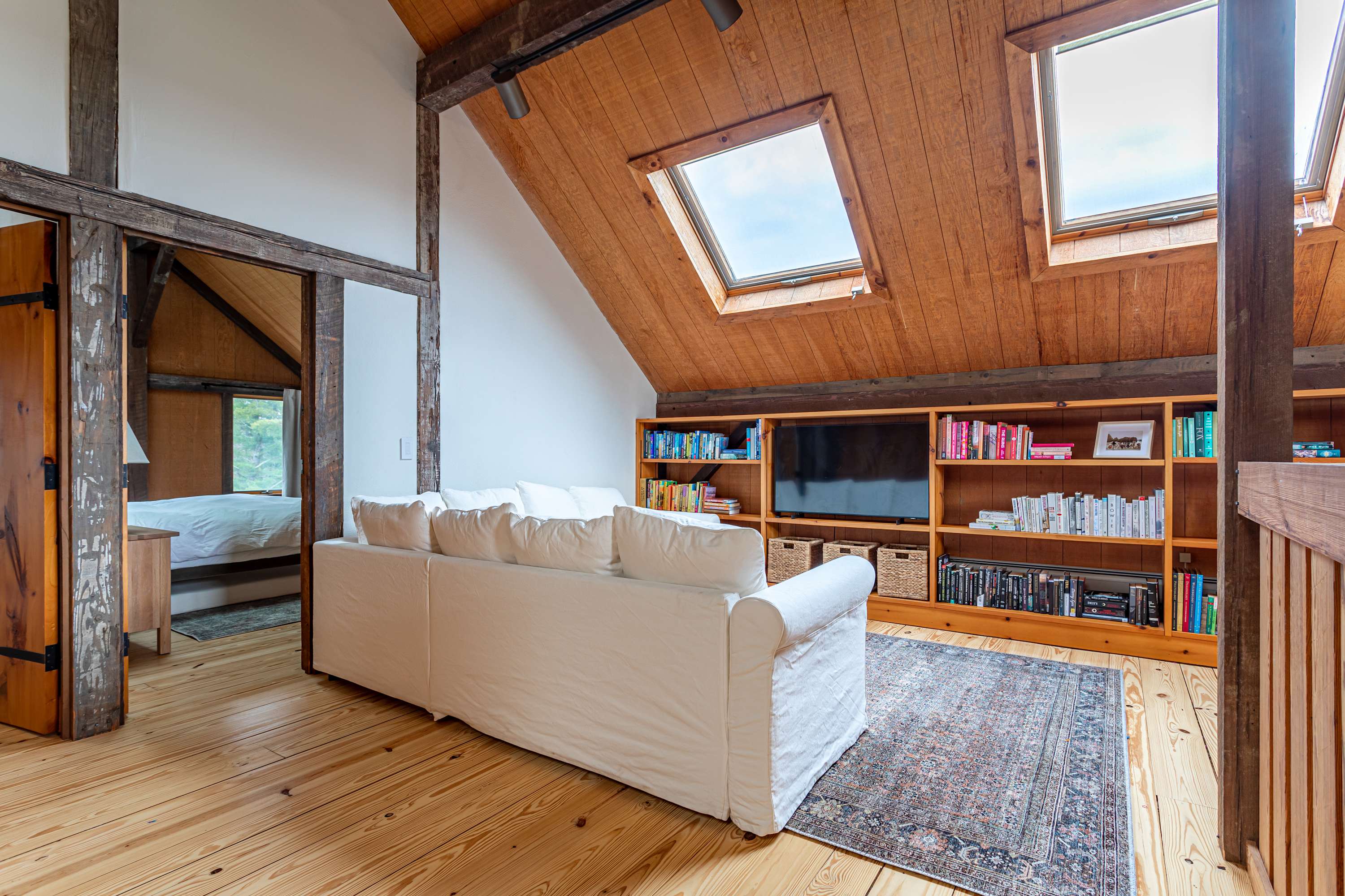 Cozy loft living area featuring a white sofa, built-in bookshelves with a television, and two bright skylights. The space overlooks the lower level and has warm wooden floors and ceilings.
