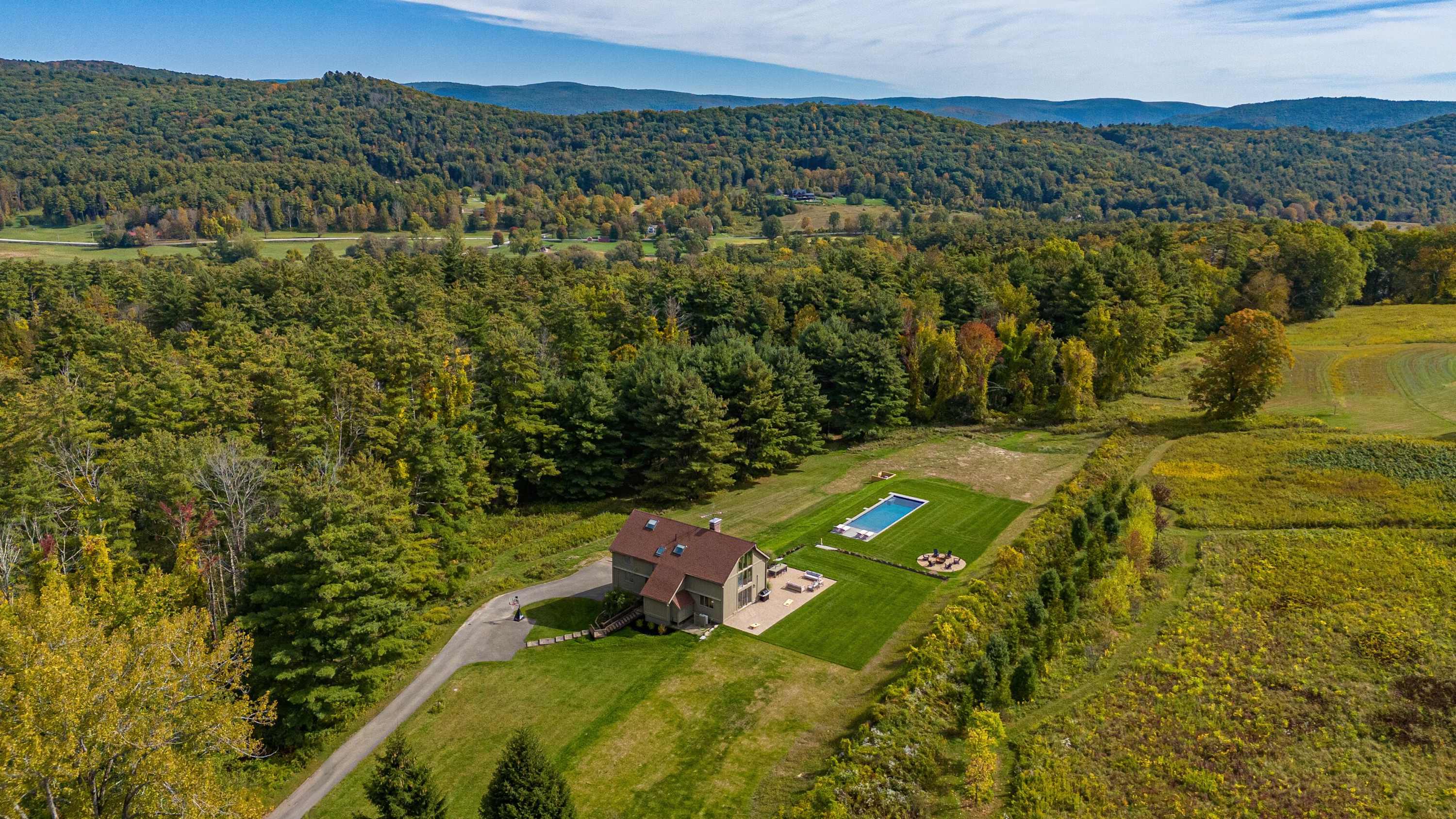 Expansive aerial view of the property, showcasing the house, pool, and vast green lawns, nestled amidst dense forests and rolling hills under a clear sky, highlighting its secluded natural setting.
