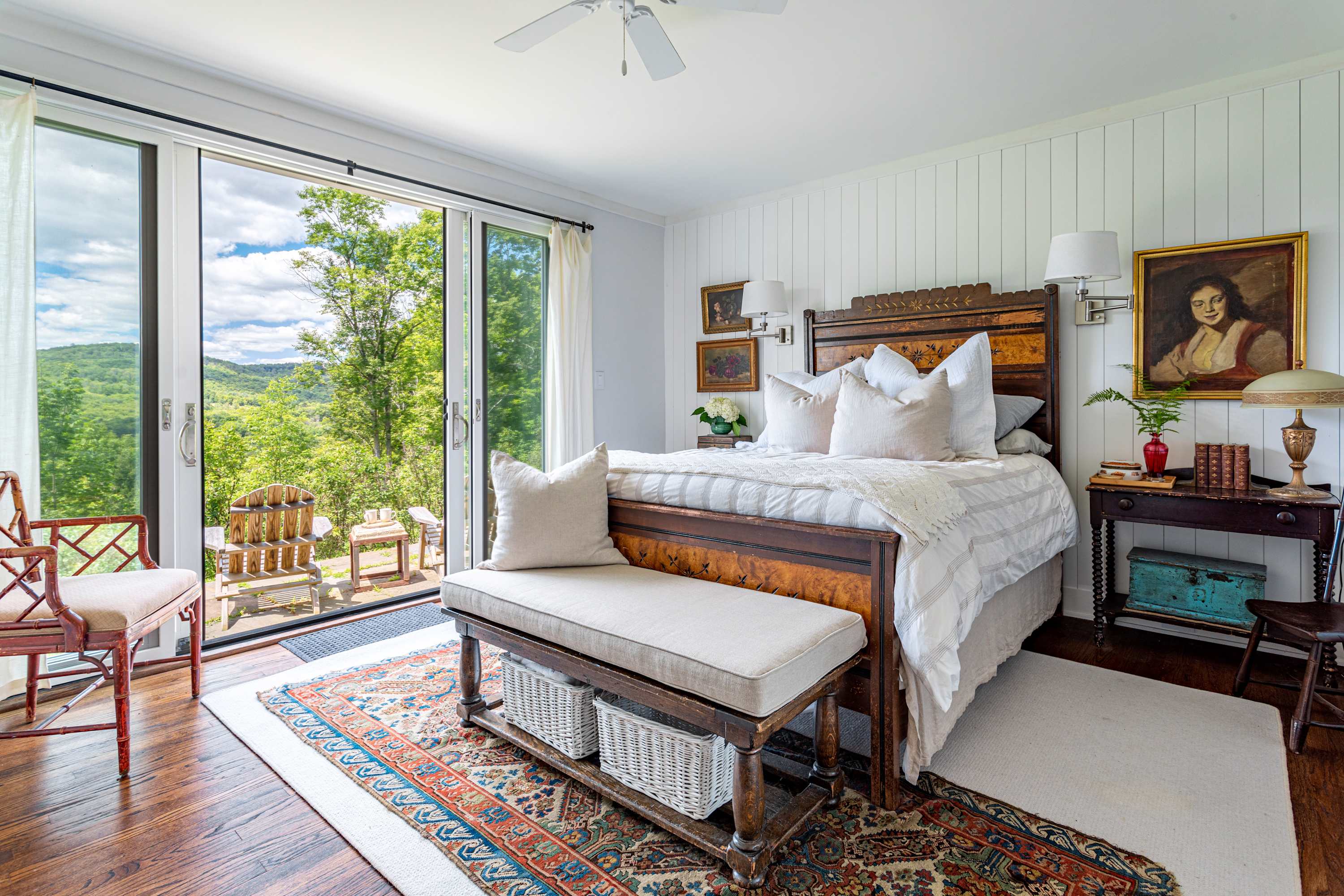 Spacious bedroom-1 with a dark wood bed frame, white bedding, and a bench at the foot of the bed, featuring large sliding glass doors opening to a deck with panoramic mountain views. The room has rich hardwood floors and white vertical paneled walls.
