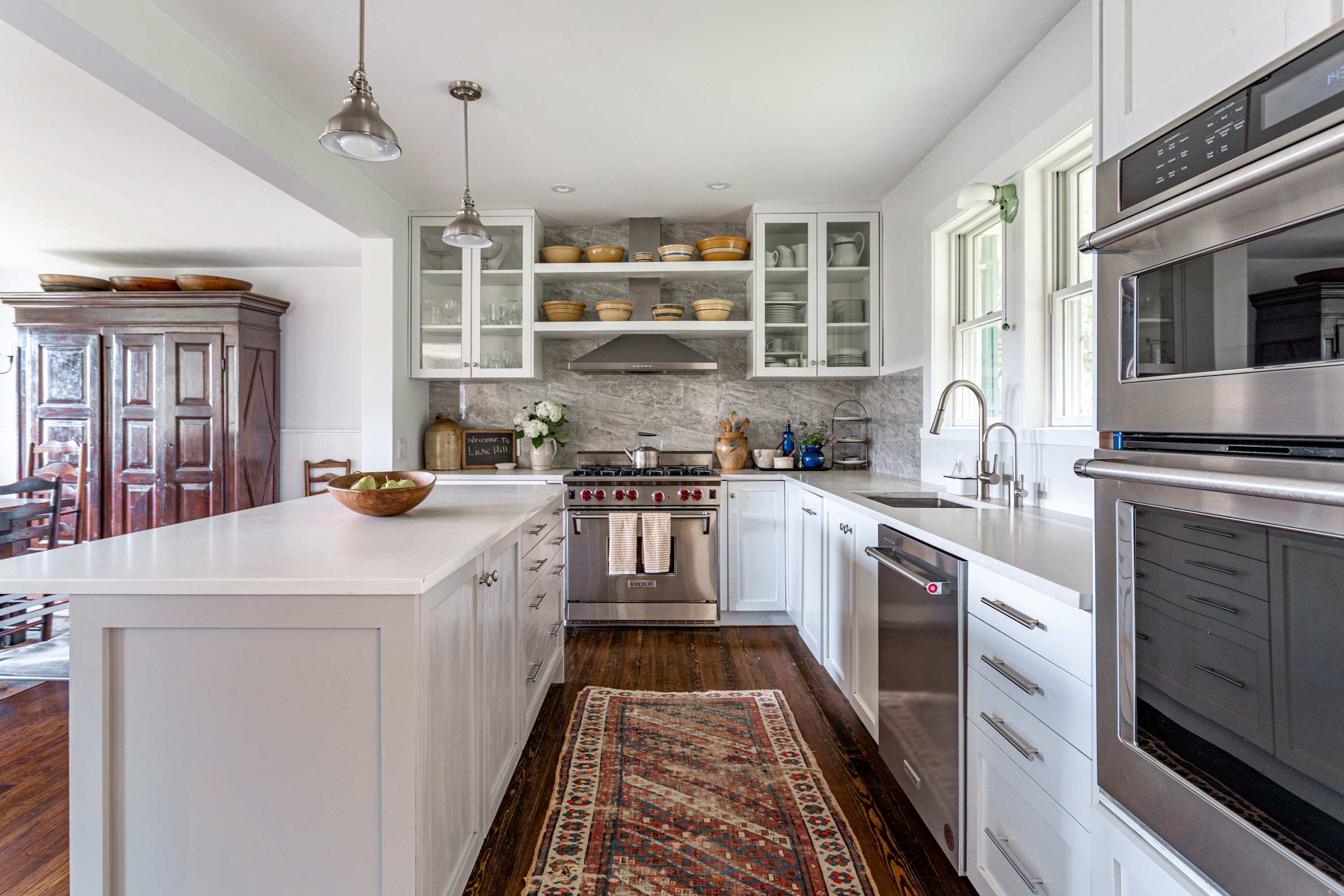 A bright, modern kitchen features white shaker cabinets, a large island, stainless steel appliances, marble backsplash, and dark hardwood floors with a colorful runner rug.
