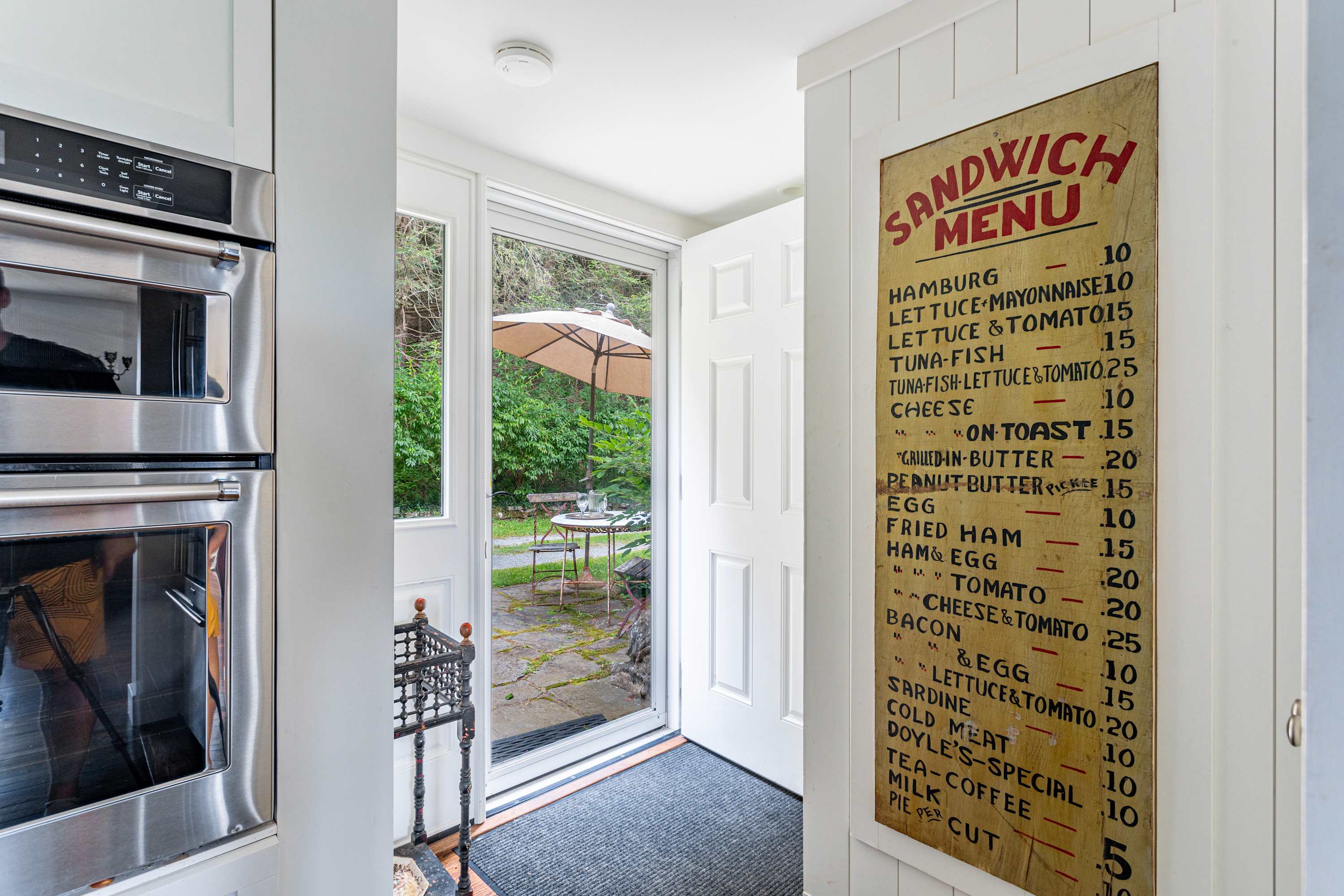 Bright entryway with white shiplap walls, featuring a vintage-style 'Sandwich Menu' sign, and a glass-paneled door leading to an outdoor patio area, with modern built-in ovens visible to the left.