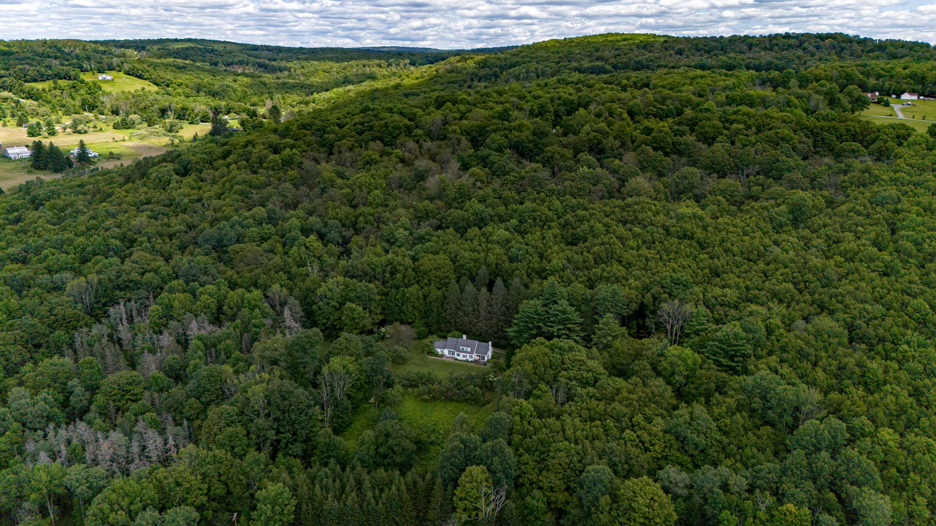 An aerial view showcases a white house with a dark roof, nestled amidst a vast, dense green forest, surrounded by rolling hills under a cloudy sky, highlighting its secluded and private location.