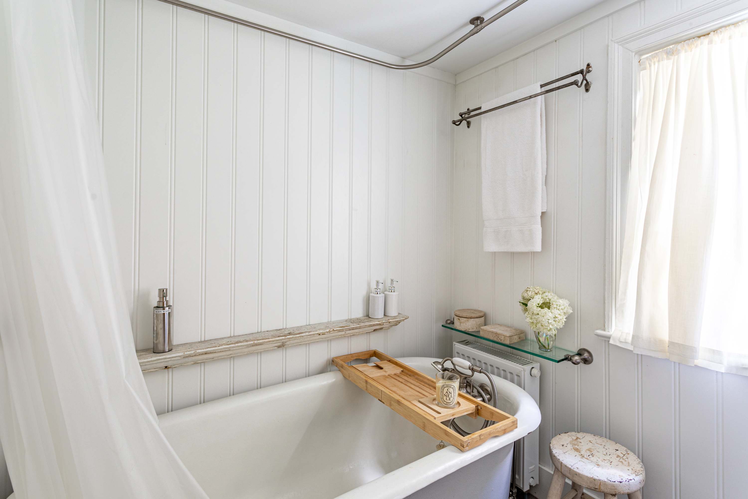 Bright white bathroom with vertical shiplap walls, a classic clawfoot tub with a wooden caddy, and a sheer white shower curtain. A small wooden shelf holds toiletries.