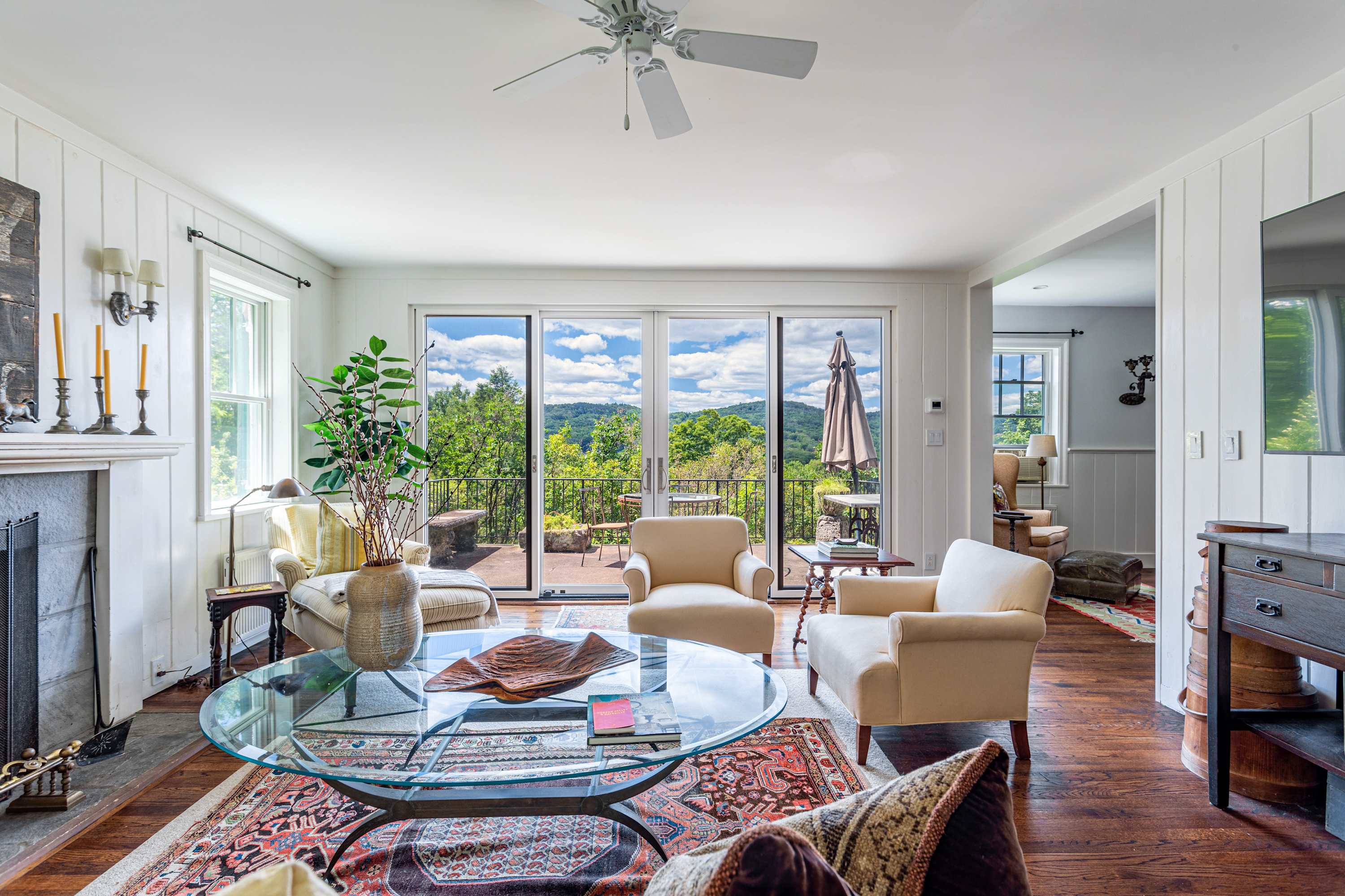 Spacious living room with warm hardwood floors, a ceiling fan, and large sliding glass doors that open to a deck, revealing expansive mountain views, furnished with comfortable cream armchairs and a glass coffee table.