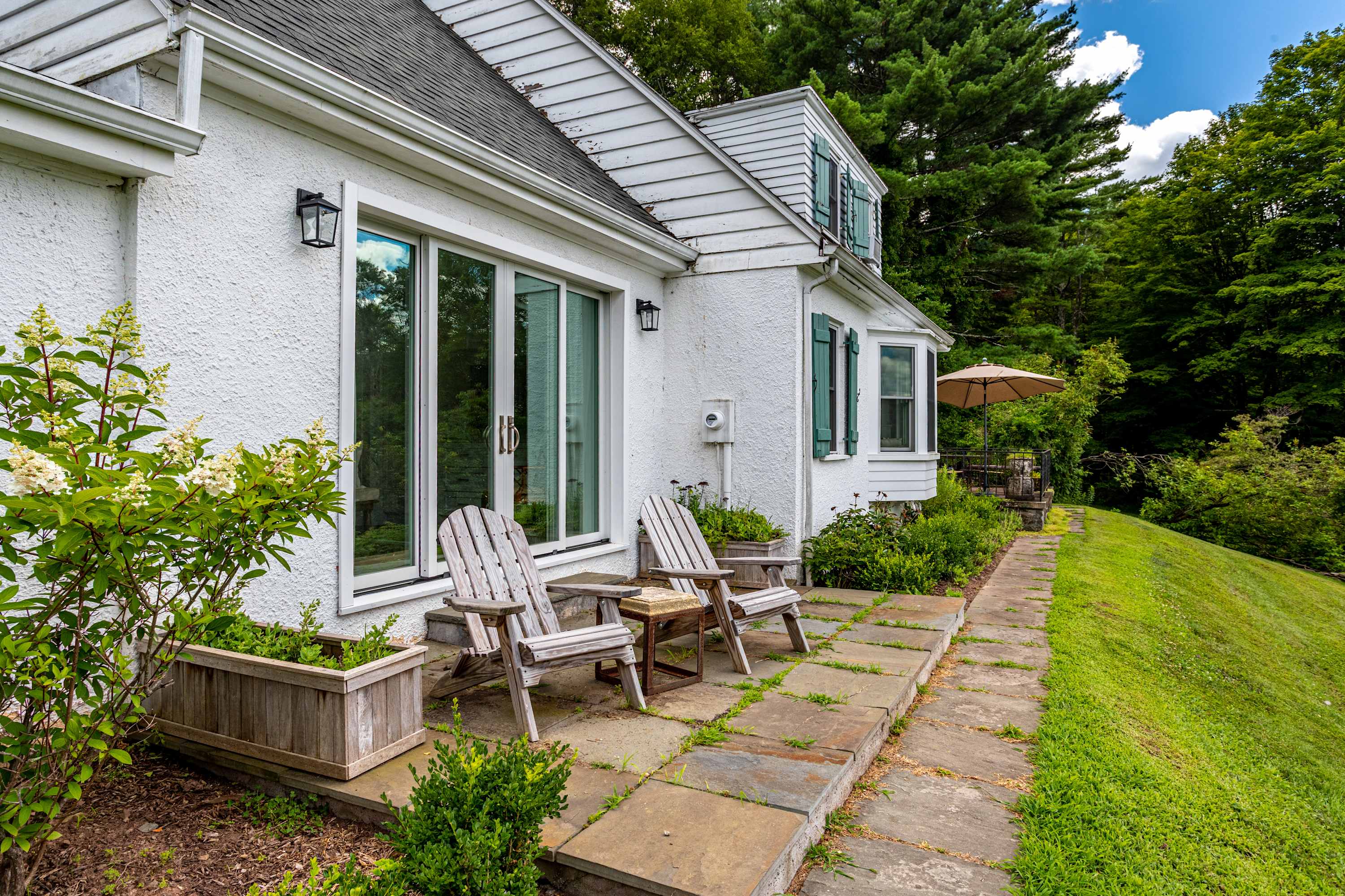 Stone patio area with two wooden Adirondack chairs and a small side table, set against the white exterior of the house with large glass sliding doors. Lush greenery surrounds the space.