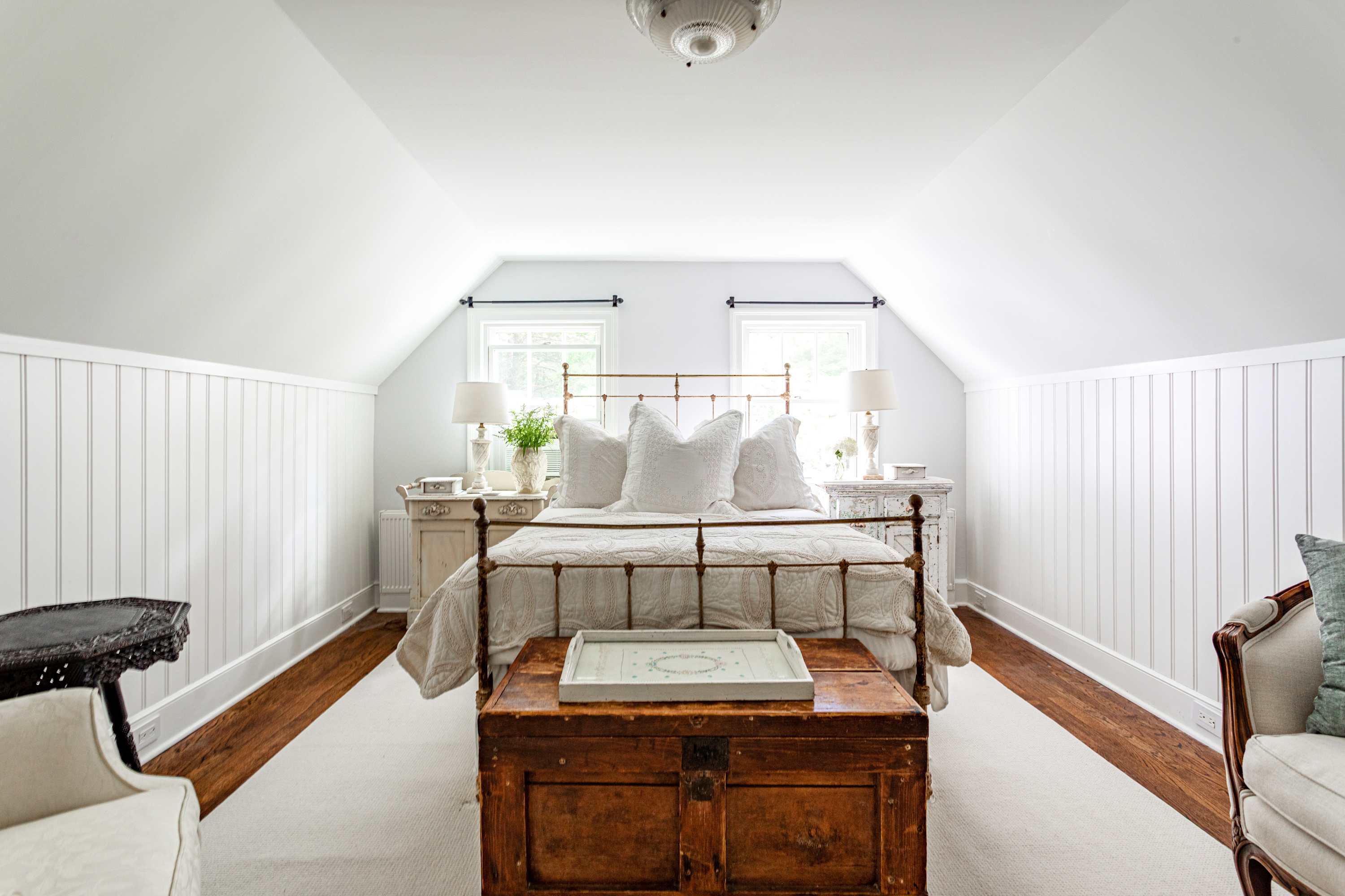 Cozy attic bedroom featuring a brass bed with white bedding, flanked by two distressed white nightstands. Sloped ceilings and white shiplap wainscoting create a charming, bright space.