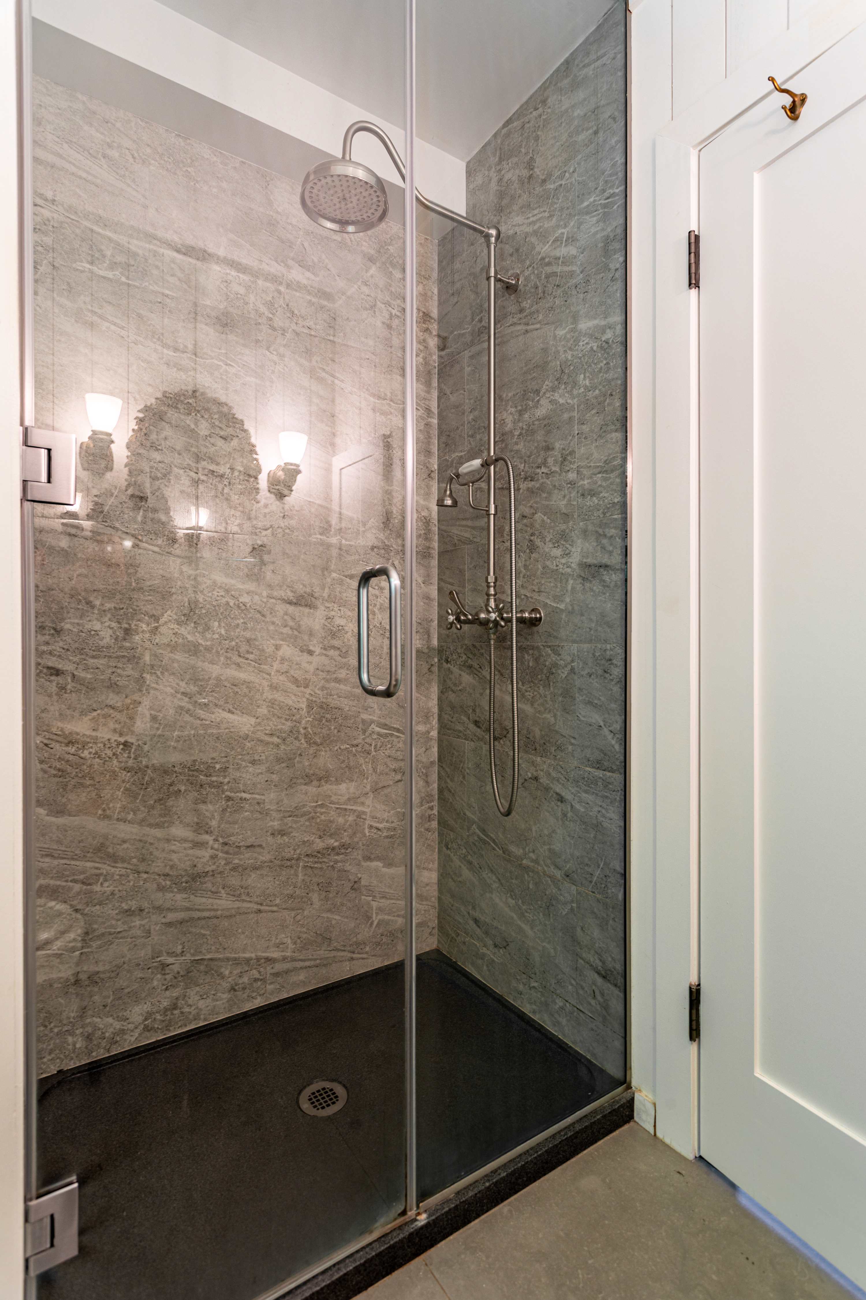 Close-up view of the modern walk-in shower from bathroom-1, showcasing grey marble-patterned tiles, a dark shower pan, and a clear glass enclosure. The shower is equipped with both a rainfall showerhead and a convenient handheld sprayer.