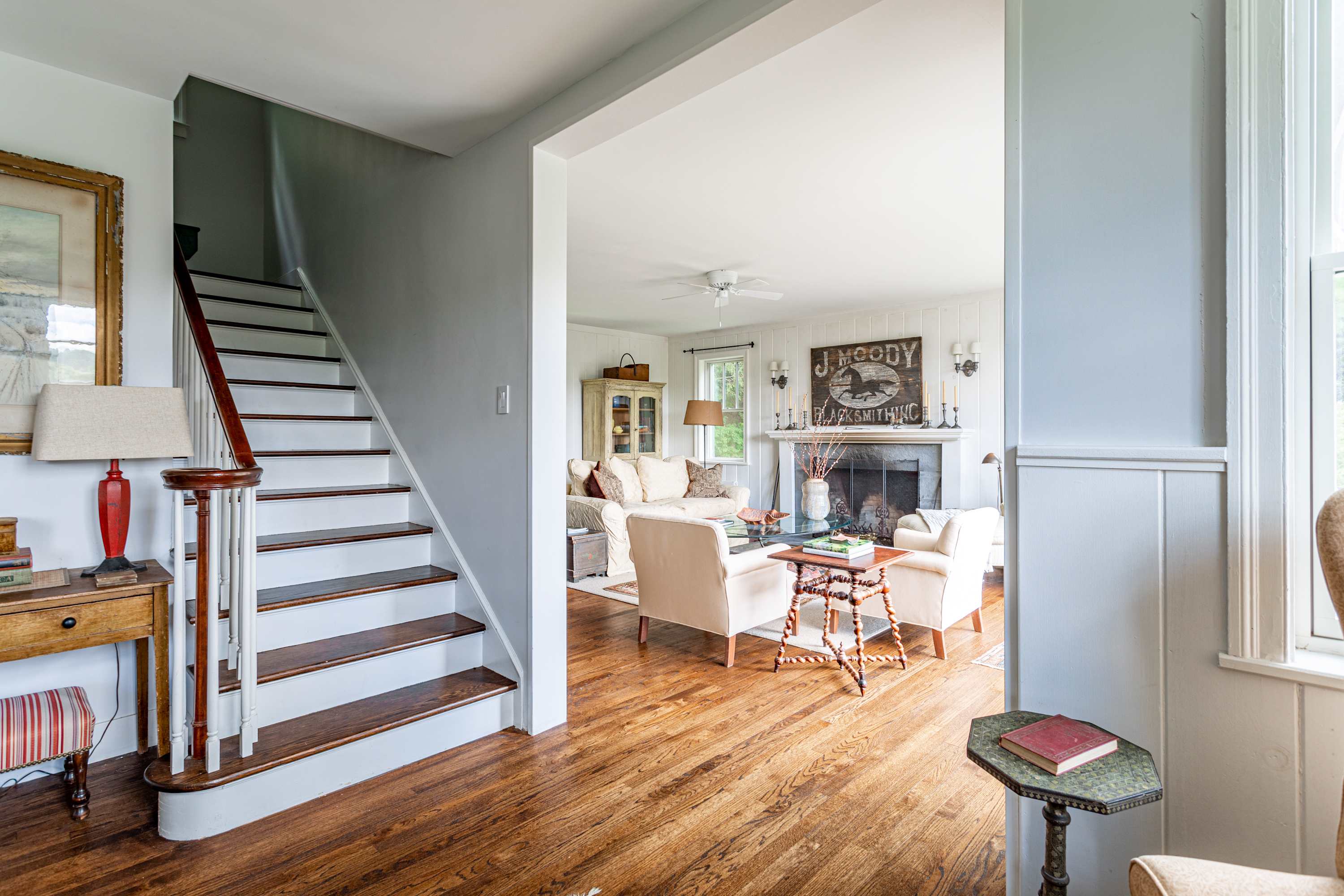 Inviting foyer with a classic wooden staircase leading upstairs, opening into a spacious living room featuring a stone fireplace with a rustic mantel, comfortable cream-colored sofas, and polished hardwood floors. Walls are light grey and white paneled.