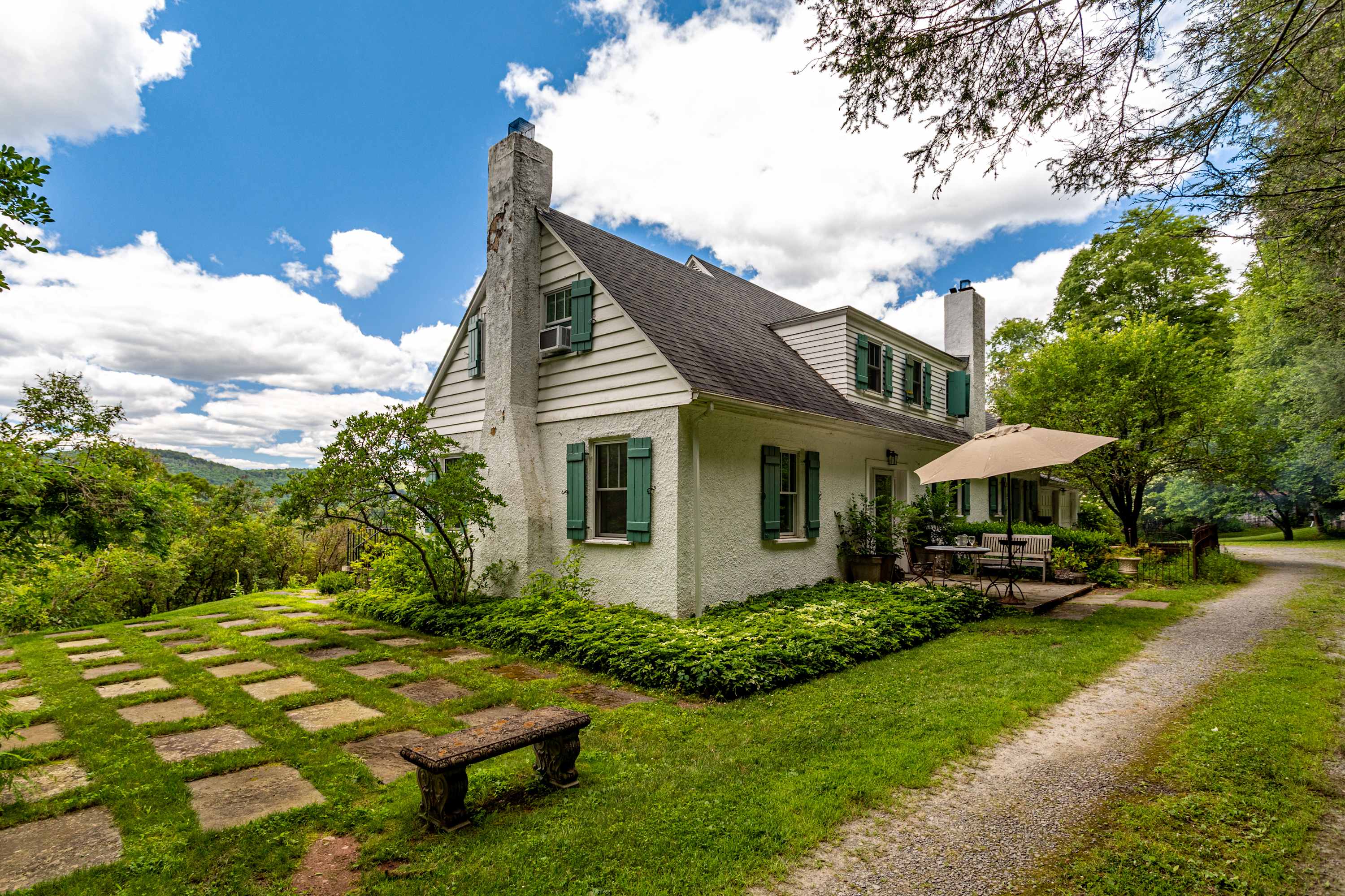 Charming white house with green shutters and multiple chimneys, surrounded by lush green grass and mature trees. A stone patio with outdoor seating and an umbrella is visible on the side.