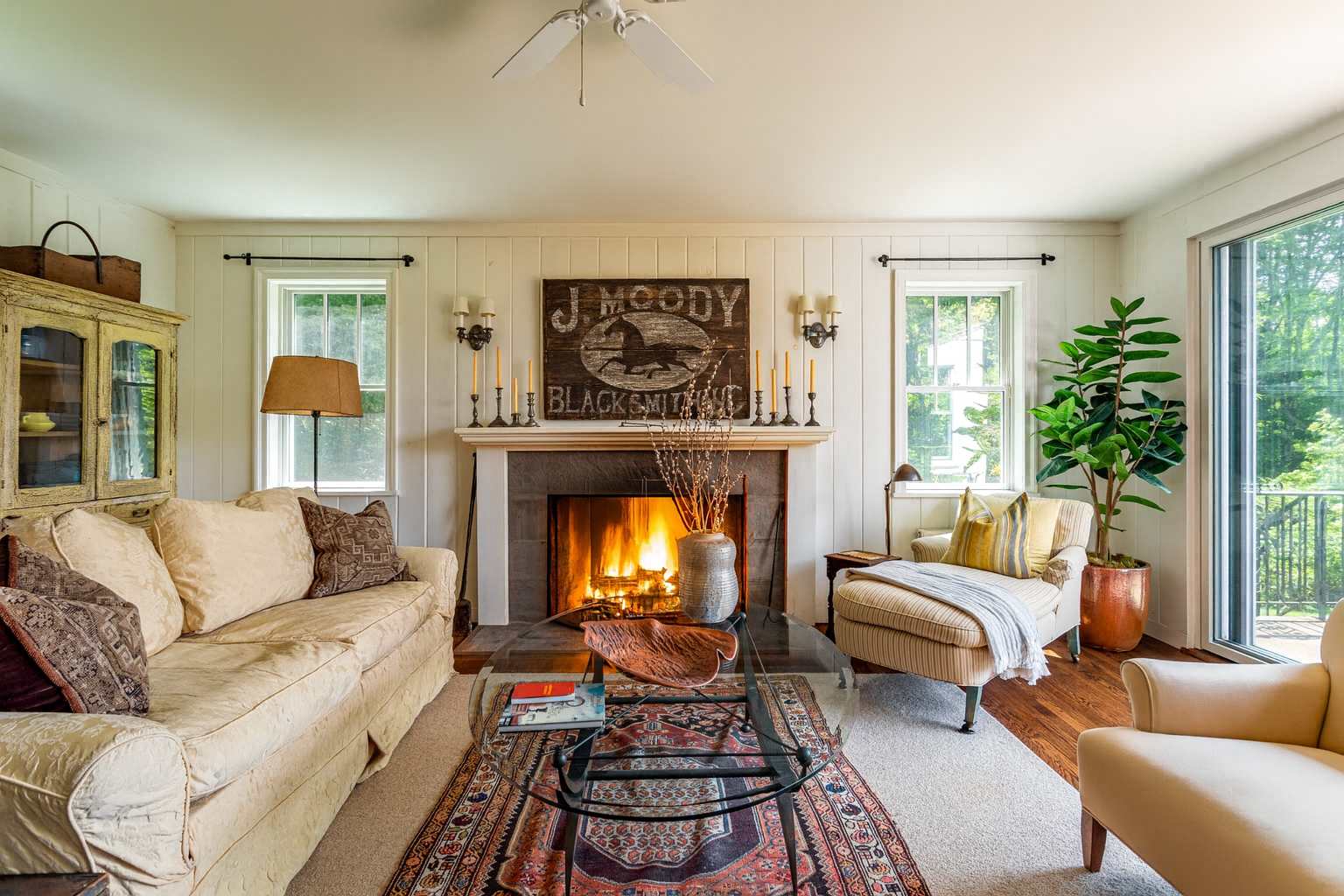 Cozy living room featuring a large stone fireplace with a roaring fire, white shiplap walls, a cream sofa, striped armchair, and a glass coffee table on an ornate Persian rug, with natural light from windows.
