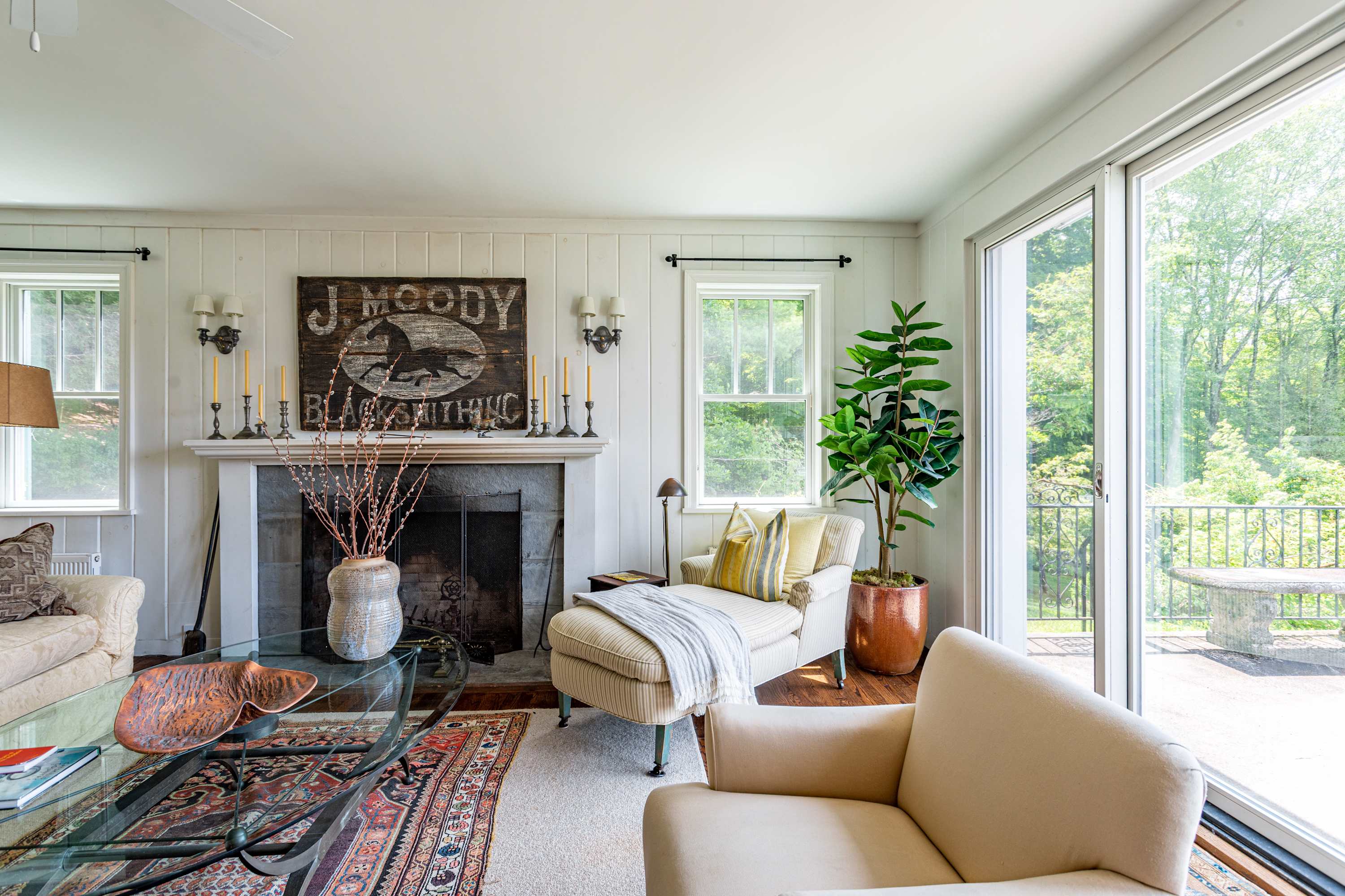 A comfortable living room centered around a stone fireplace with a white mantel, featuring a chaise lounge, glass coffee table, and a sliding door leading to an outdoor deck with mountain views.