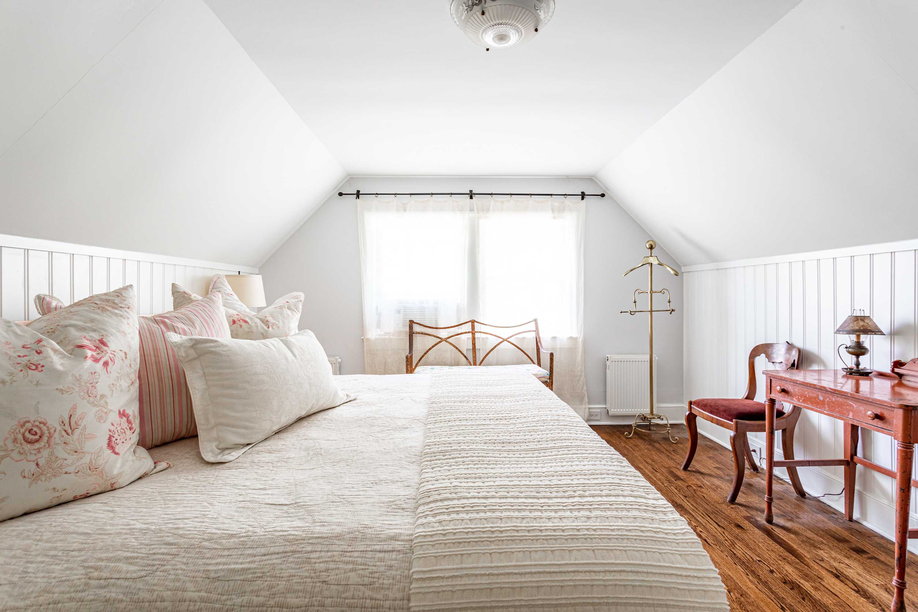 Another view of bedroom-2, highlighting the charming sloped ceilings and a bright window at the far end, with a vintage-style chair and small wooden desk visible. The room maintains its white paneled walls and warm hardwood floors.