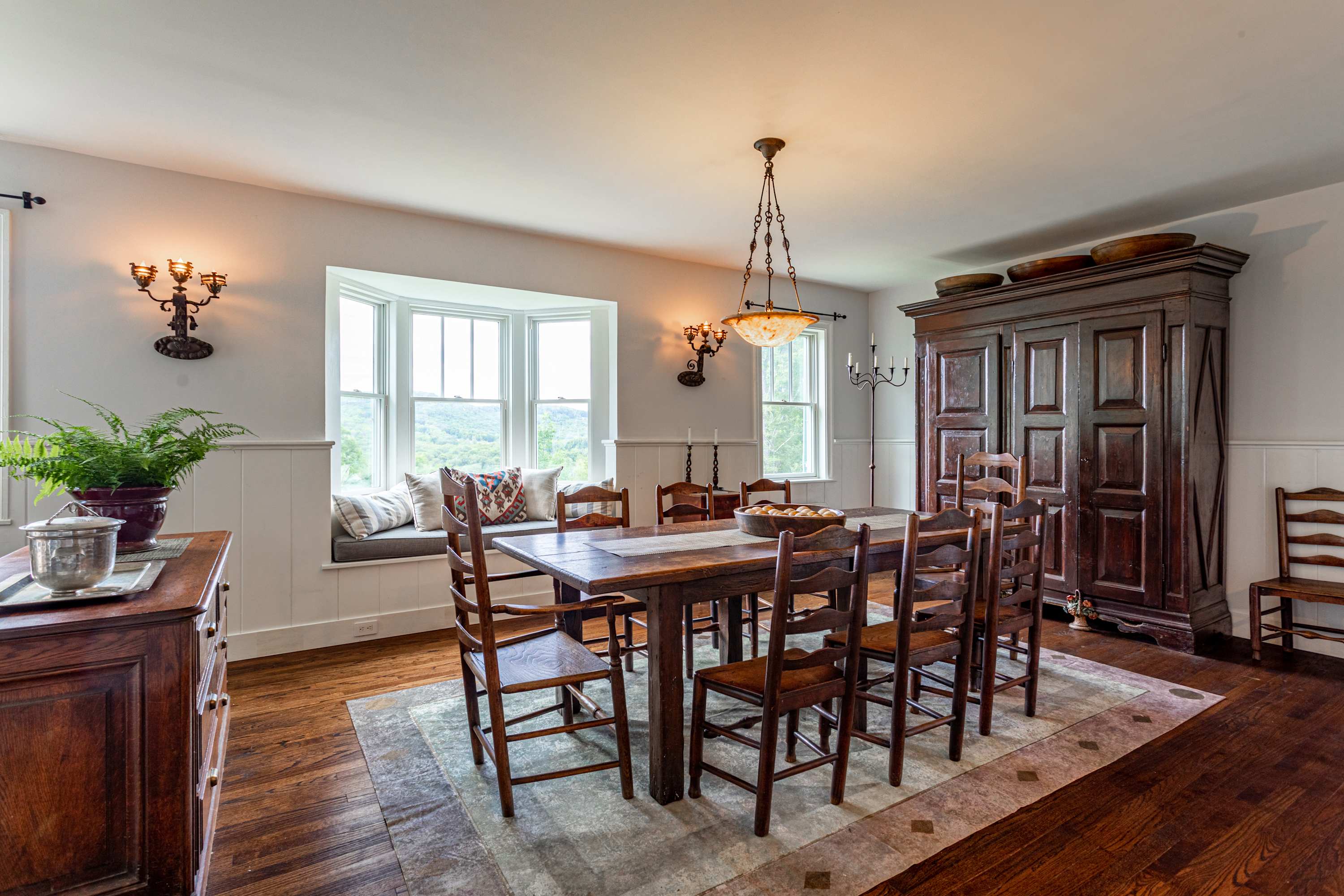 A rustic dining room features a long wooden table with matching chairs, a built-in window seat with cushions, and a large dark wood armoire, all on dark hardwood floors.