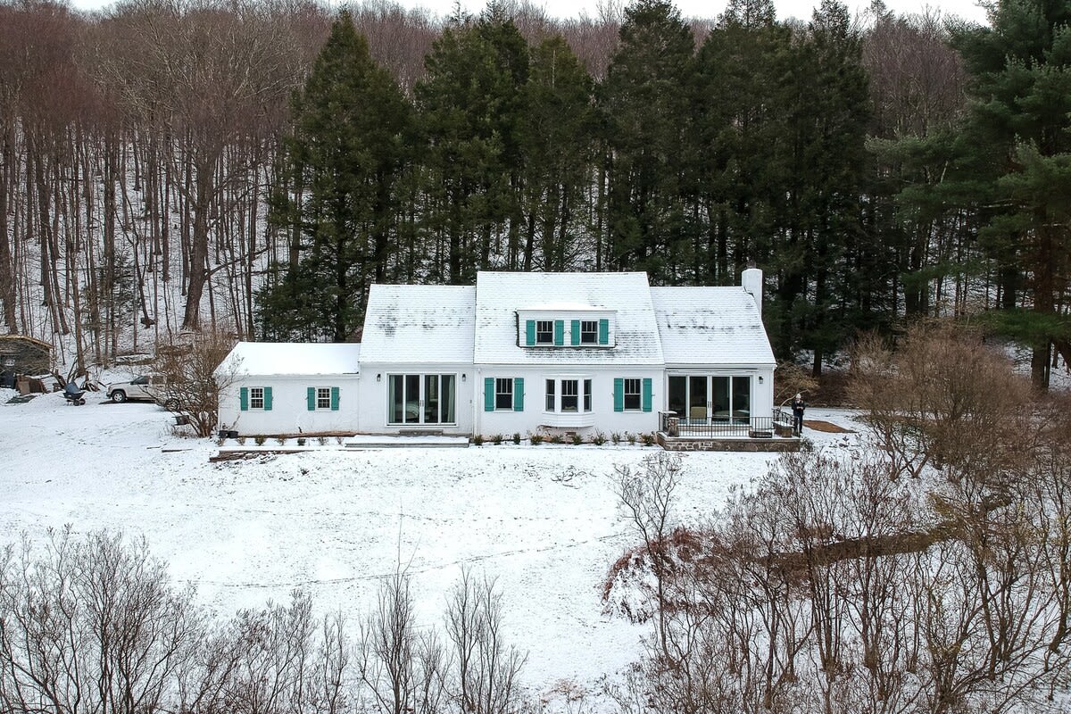 An aerial view captures the charming white house with teal shutters and a snow-covered roof, surrounded by a blanket of snow and a mix of bare deciduous trees and dark green evergreens, showcasing its winter appeal.