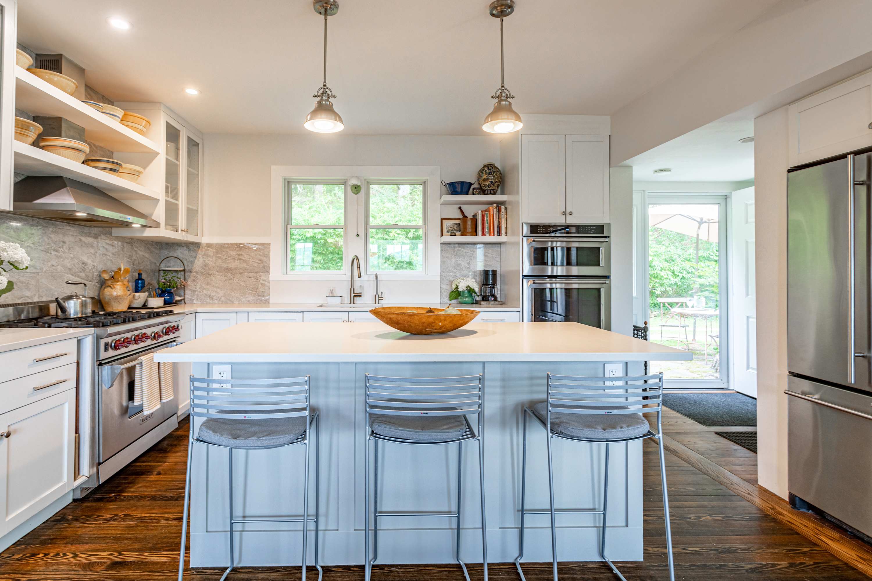 A spacious kitchen with a large white island and three bar stools, white cabinets, stainless steel appliances, and a view of the outdoors through a doorway.