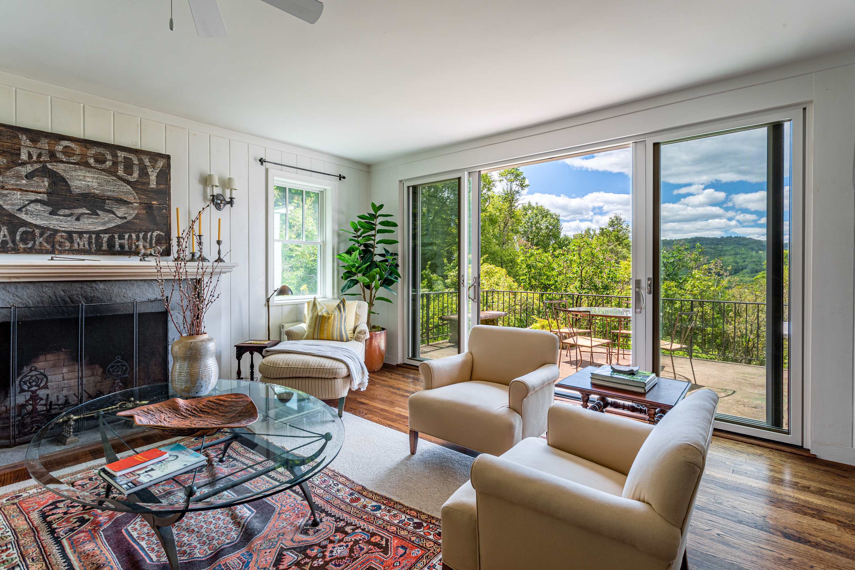 A bright living room with a stone fireplace, white paneled walls, and a large sliding glass door opening to a deck with panoramic views of lush green mountains and a blue sky.