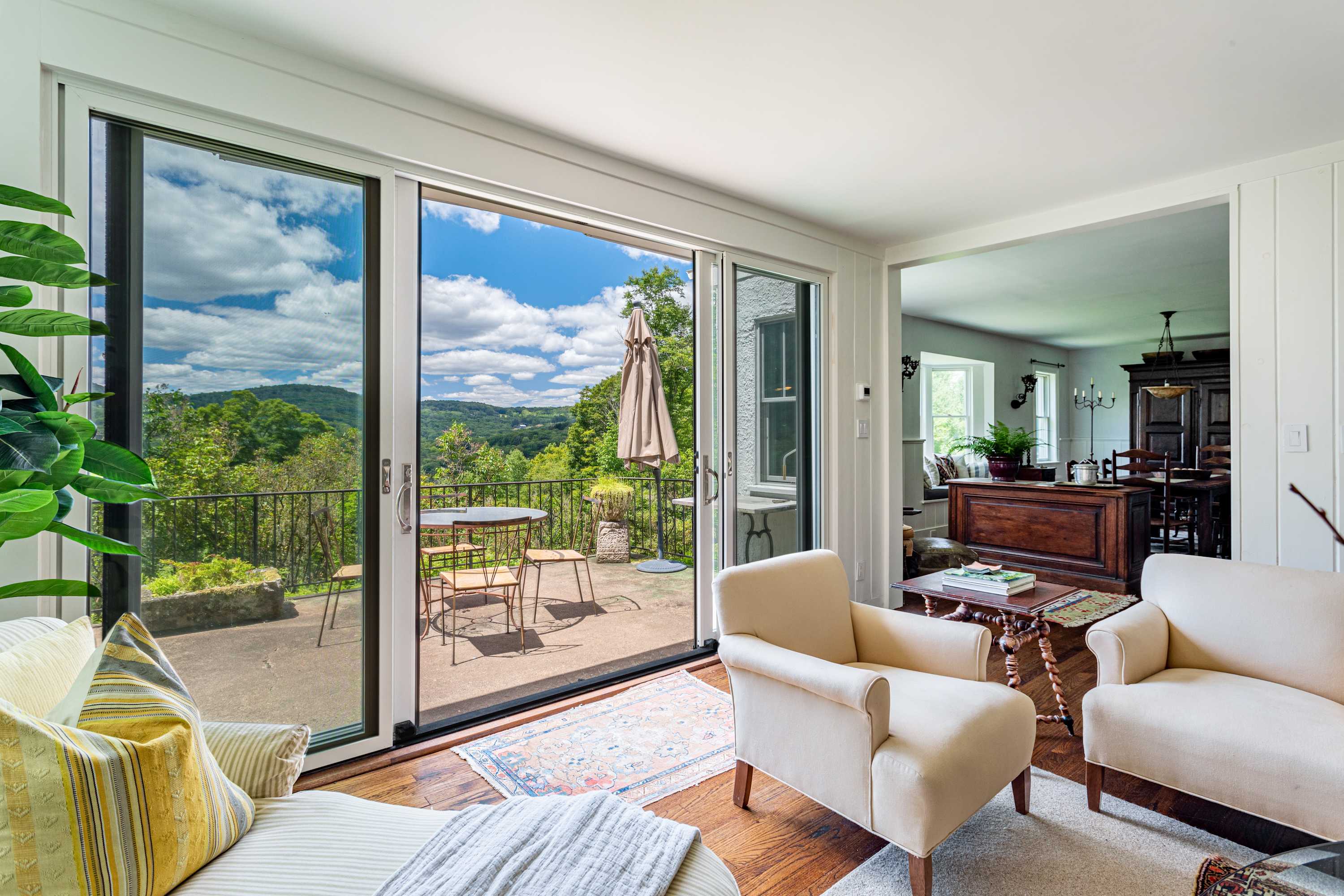 A cozy living area with light-colored armchairs and a sliding glass door opening to an outdoor patio with a table and chairs, offering expansive mountain views.