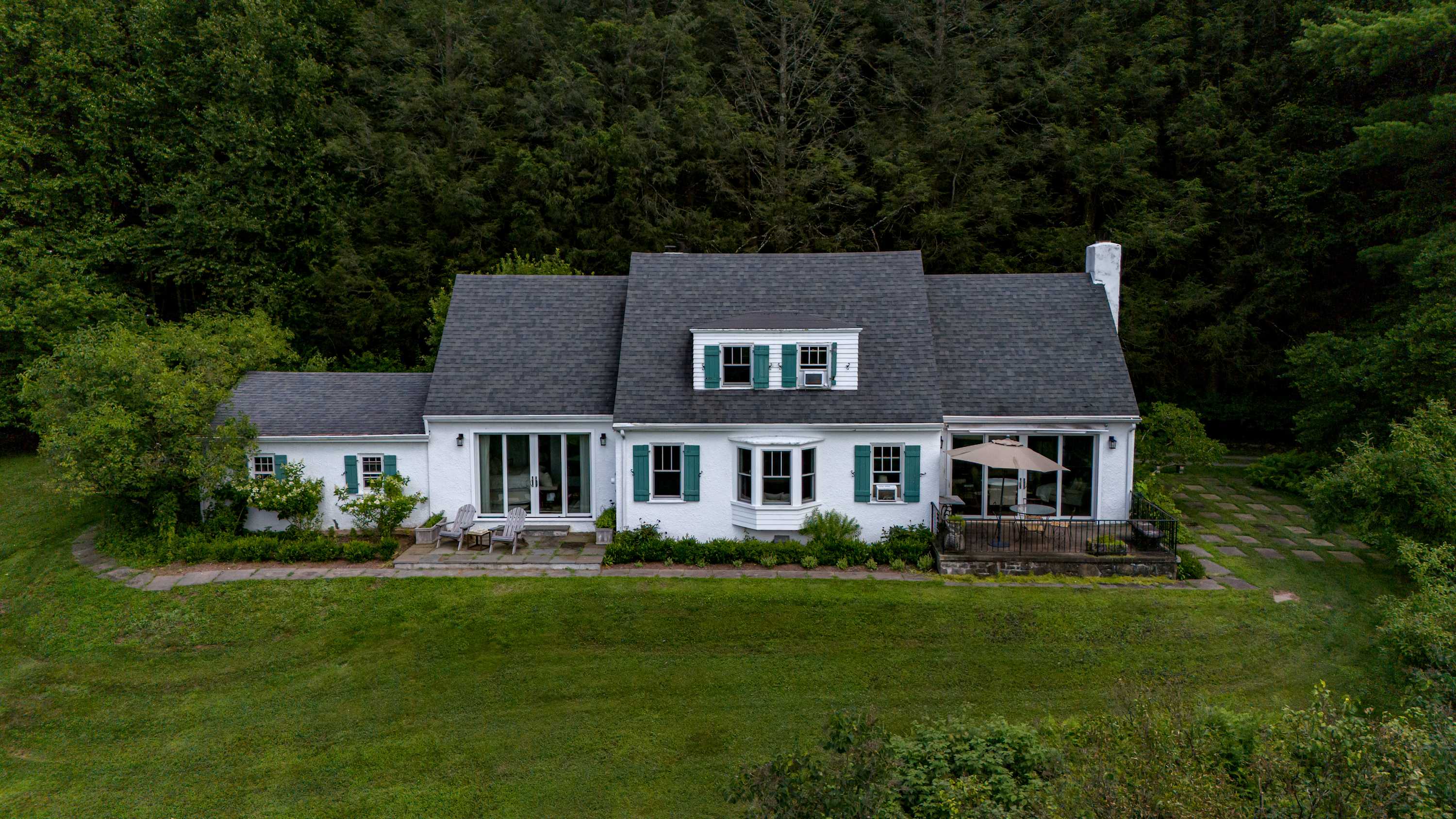Closer aerial view of the white house with a dark roof and green shutters, showcasing its layout and surrounding green lawn and trees. A patio with an umbrella is visible on the side.