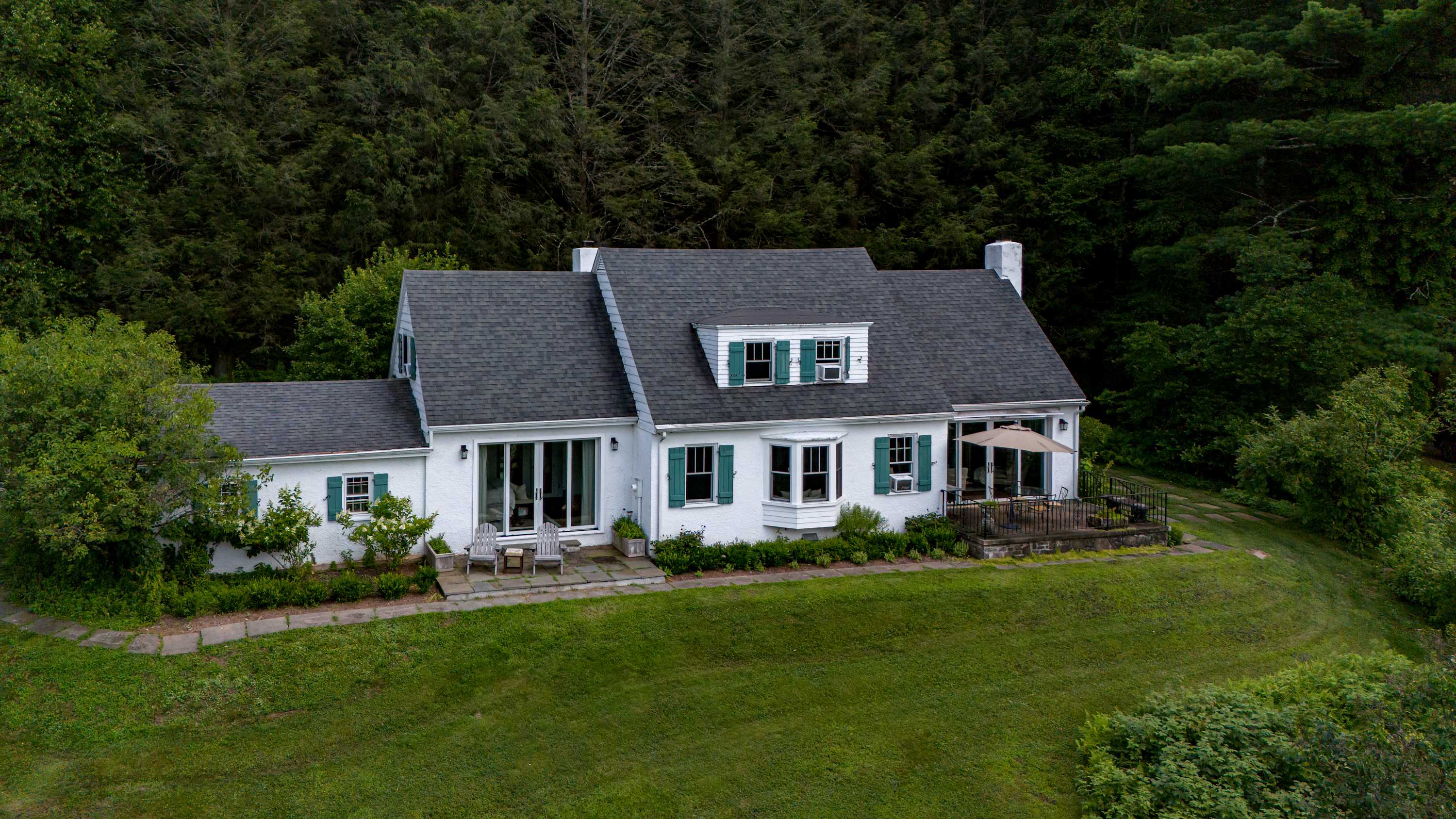 Aerial view of a charming white house with a dark gray shingle roof and green shutters, nestled against a dense forest backdrop, featuring a well-maintained grassy lawn and an outdoor patio area.