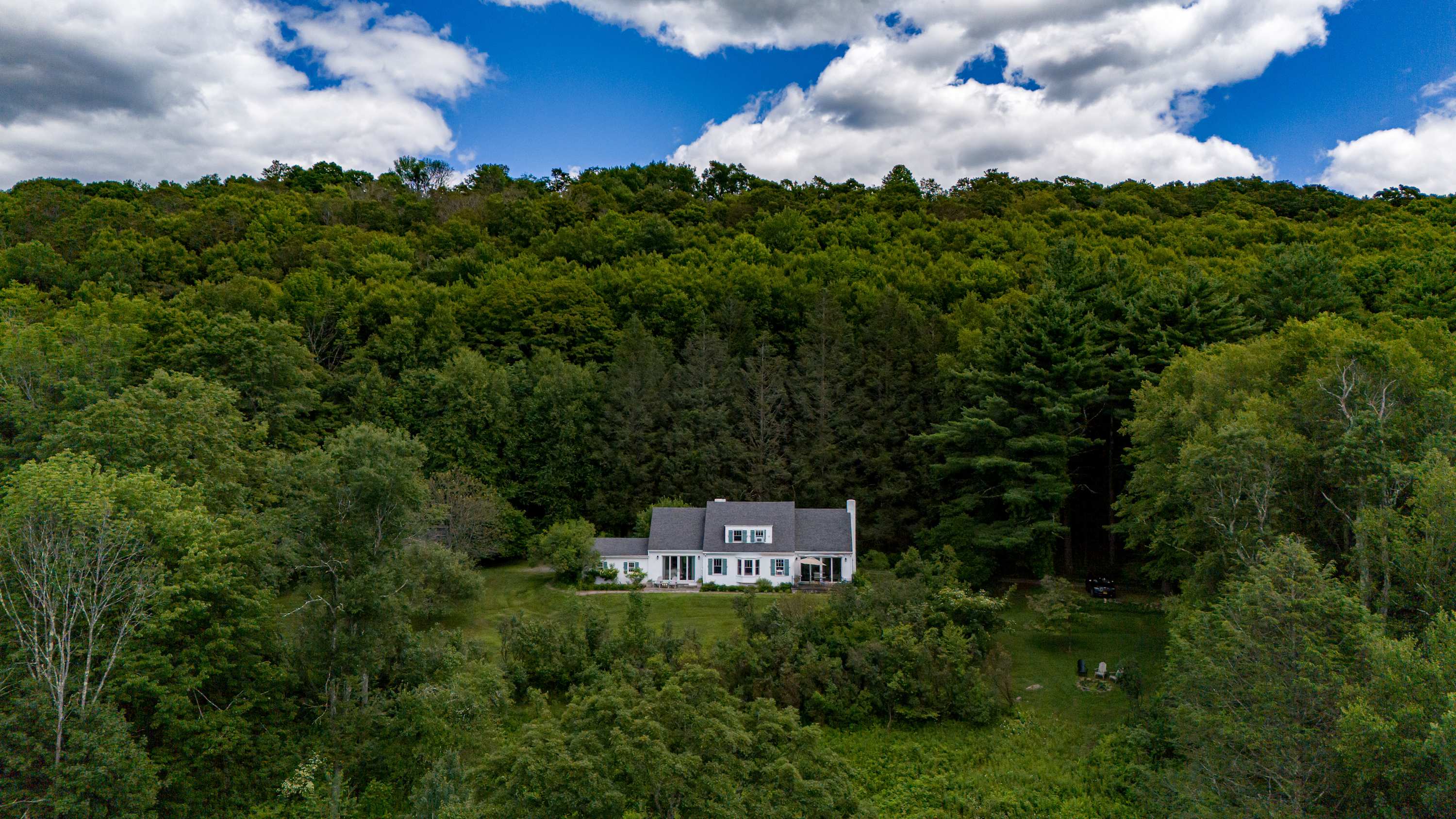 Aerial view of the white house with a dark roof and green shutters, nestled amidst a dense forest of vibrant green trees under a bright blue sky with fluffy white clouds.