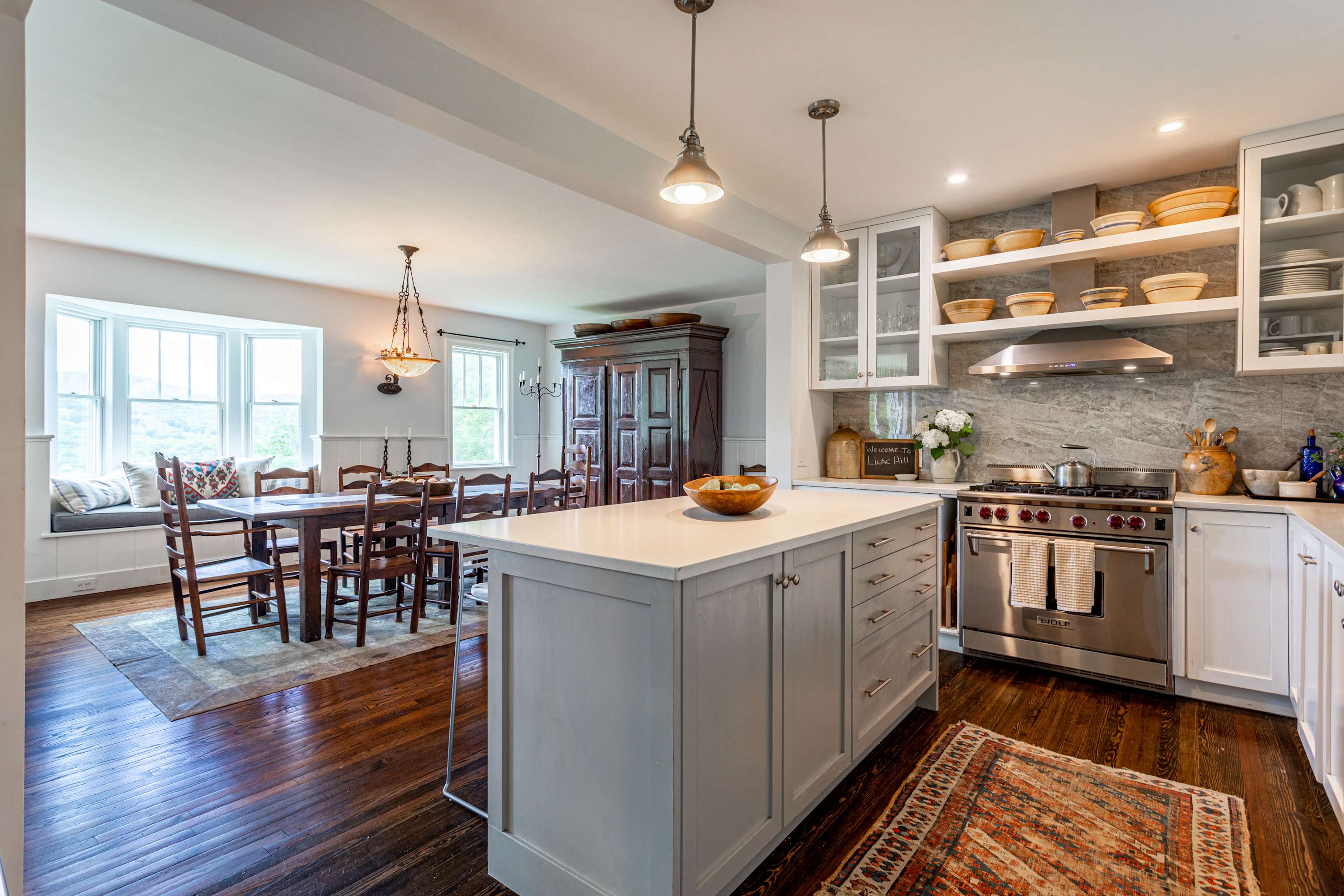 A wide view of the kitchen showing a light gray island, white perimeter cabinets, stainless steel range, and a glimpse into the adjacent dining area with a large wooden table.