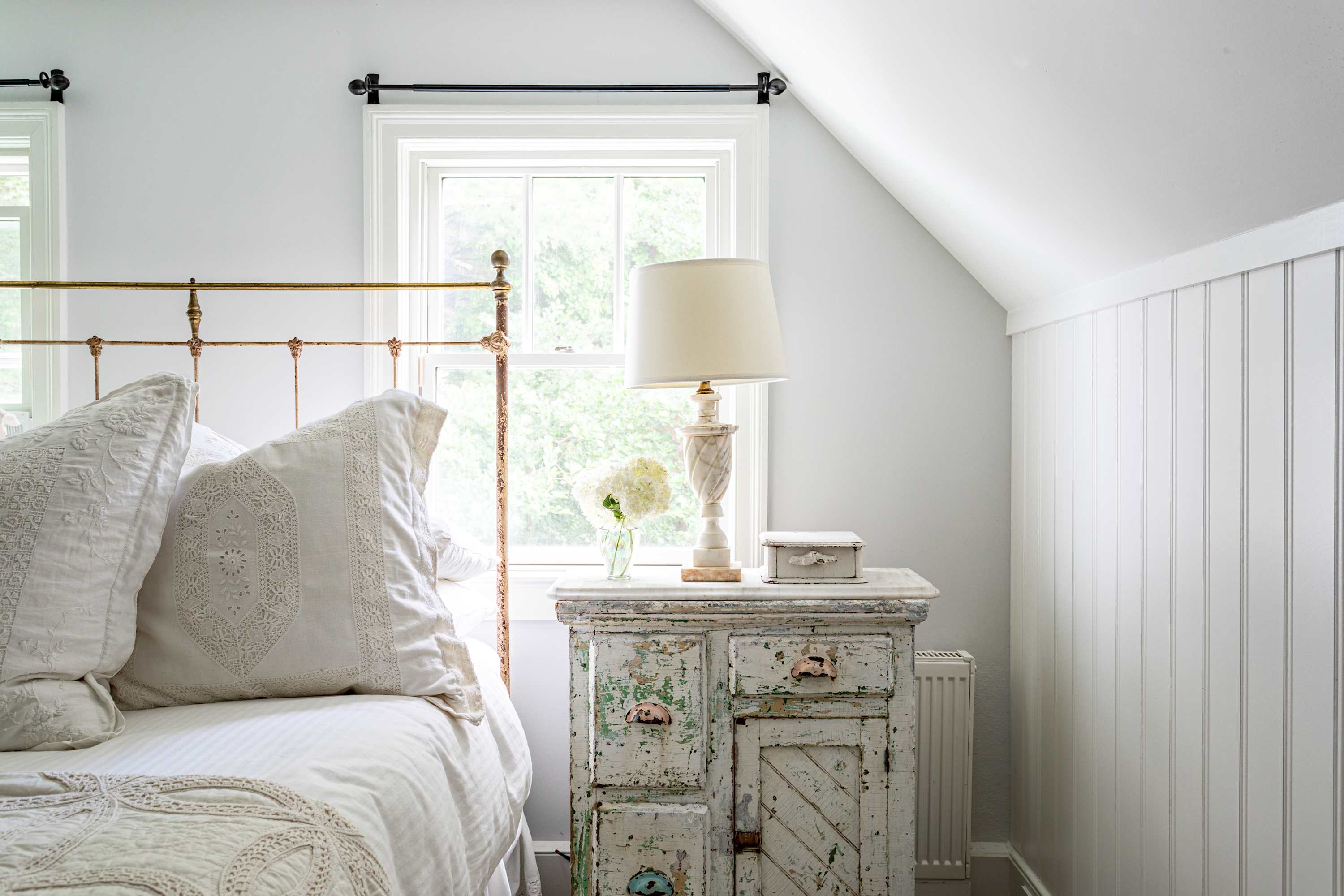 Close-up of a brass bed with white pillows and a distressed white nightstand, featuring a decorative lamp and a vase of white hydrangeas. A window provides natural light.