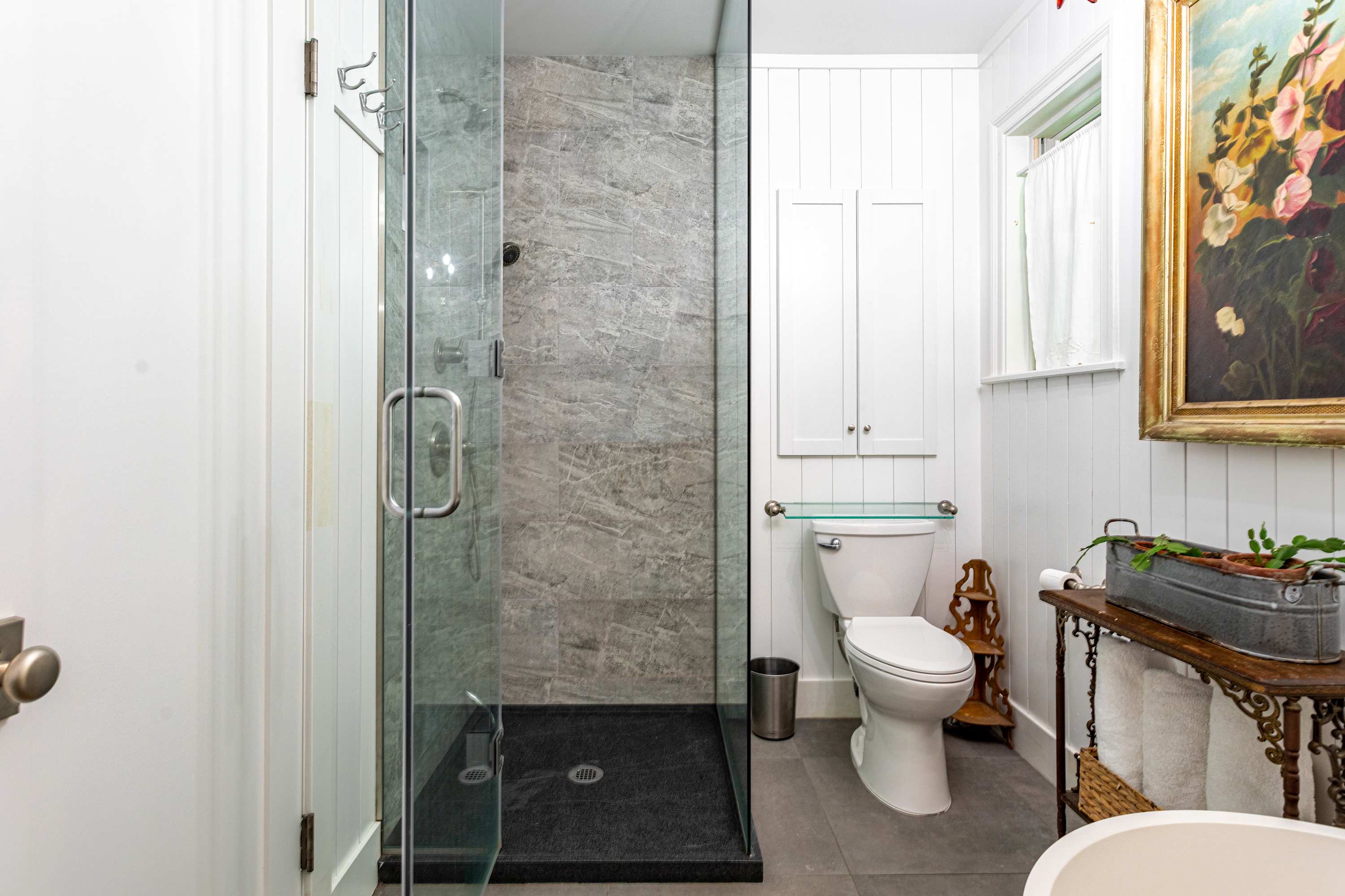 Modern bathroom-1 featuring a sleek glass-enclosed shower with grey marble-patterned tiles and a dark shower pan, alongside a white toilet and white vertical paneled walls. A small window provides natural light to the space.