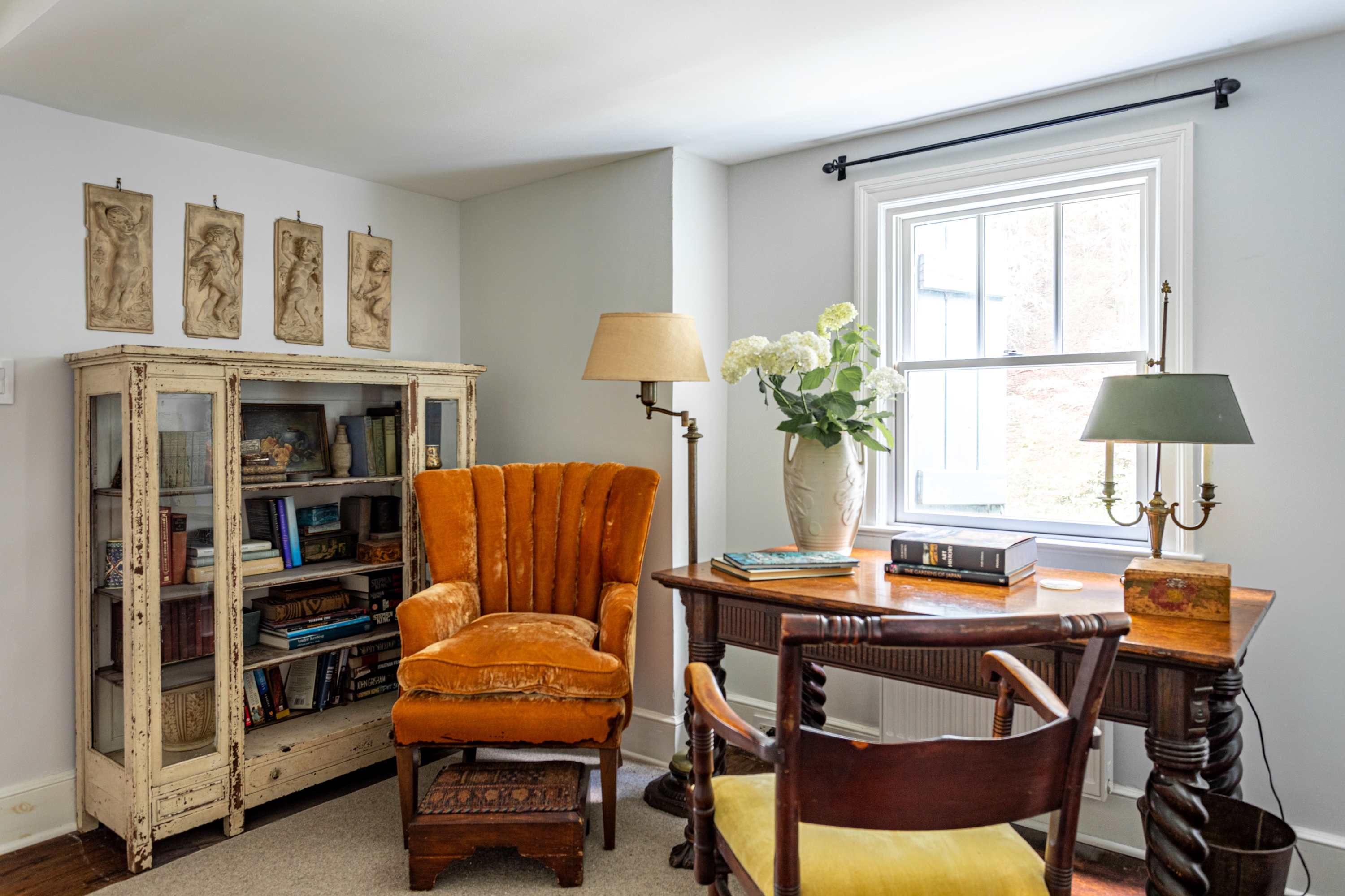 Inviting office space with a vintage wooden desk, an orange velvet armchair, and a glass-fronted display cabinet filled with books. White walls and a window create a bright, functional area.