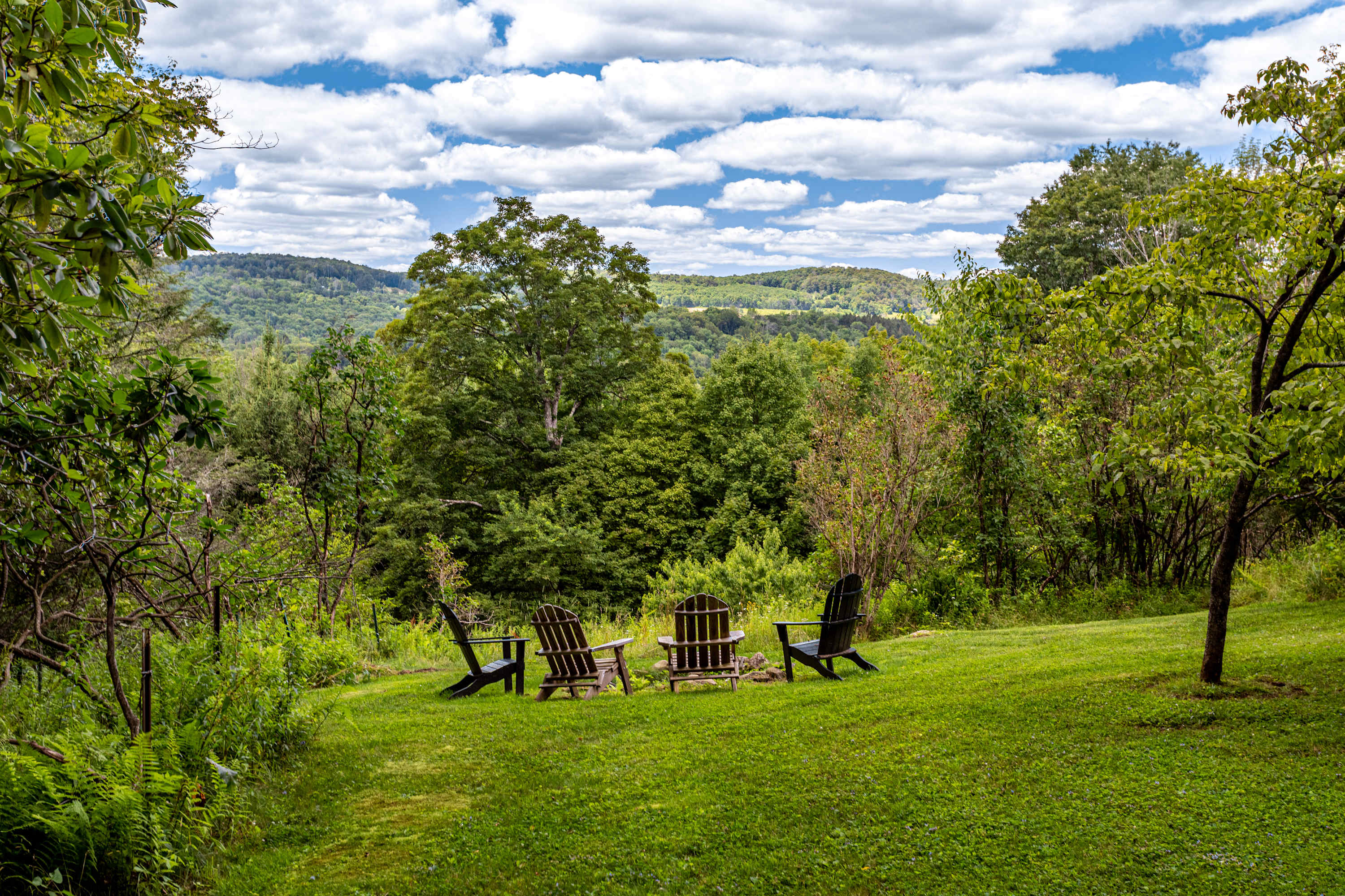 Expansive green lawn with four dark Adirondack chairs arranged in a circle, surrounded by lush trees and offering a serene distant view of rolling green mountains under a partly cloudy sky.