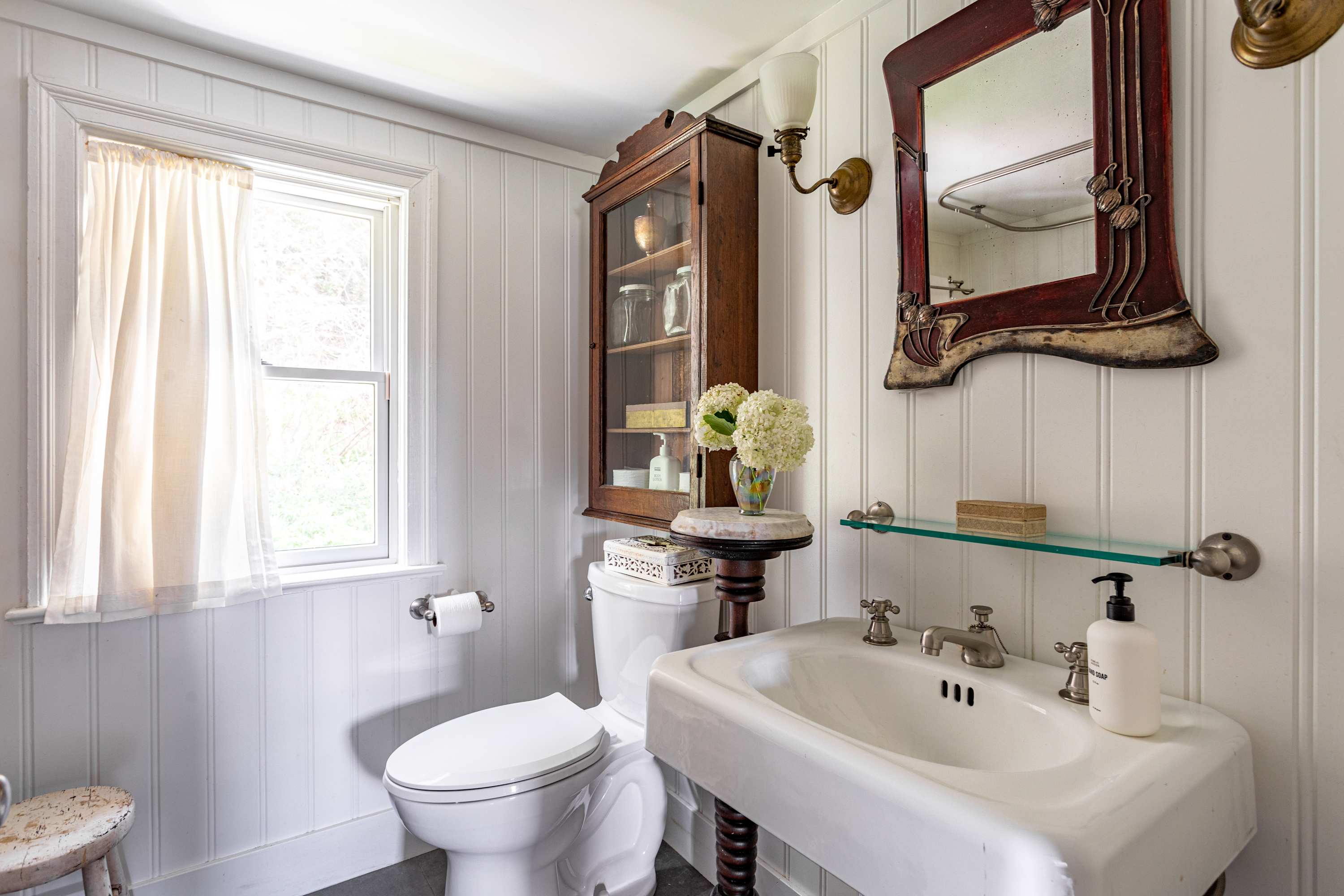 White bathroom with vertical shiplap walls, a vintage pedestal sink, and a toilet. A dark wood display cabinet and an ornate mirror add a touch of antique charm.