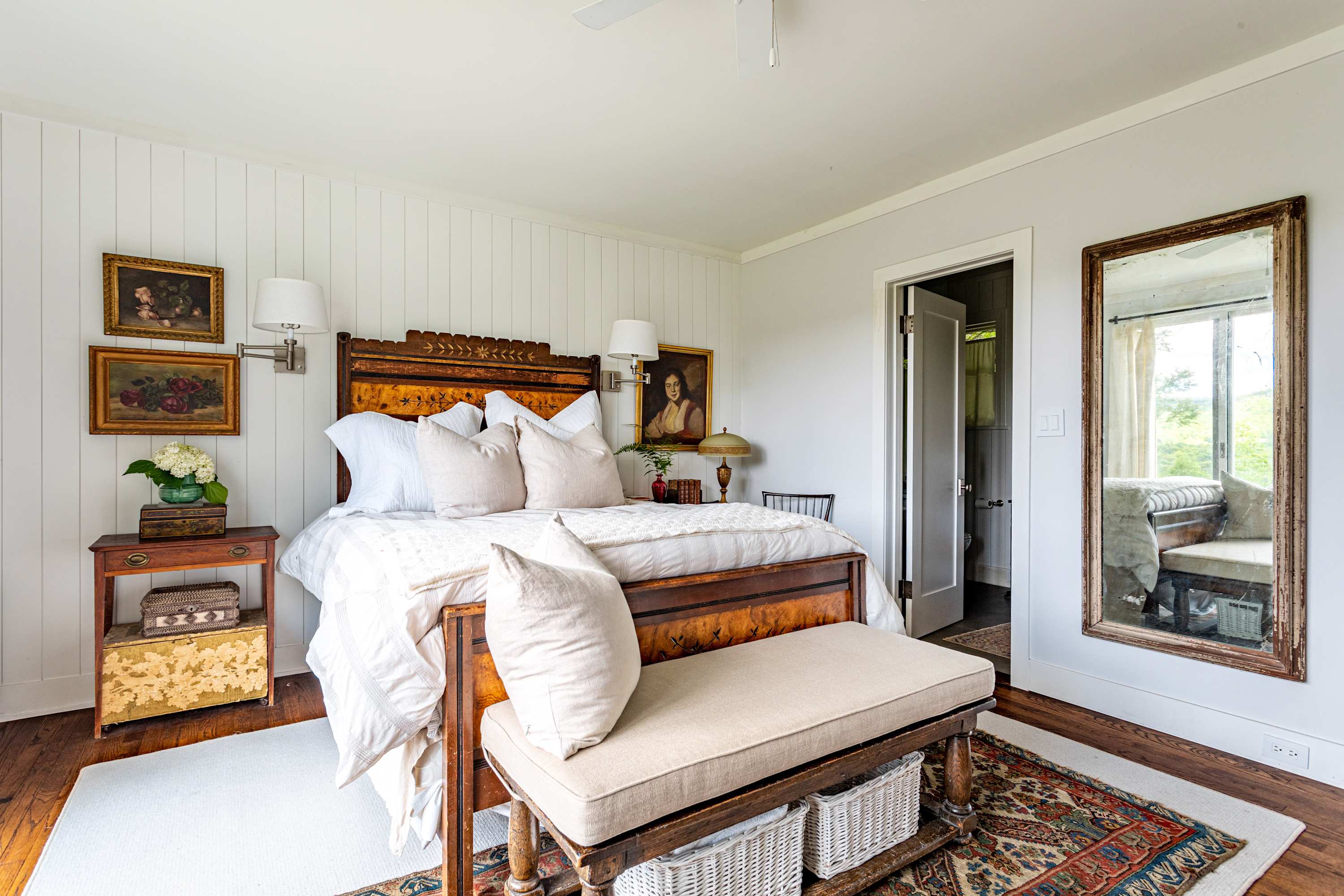 Another perspective of bedroom-1, showcasing the ornate dark wood bed, white bedding, and a large rustic mirror leaning against a white paneled wall, with a doorway leading to an adjacent room. The room features polished hardwood floors.