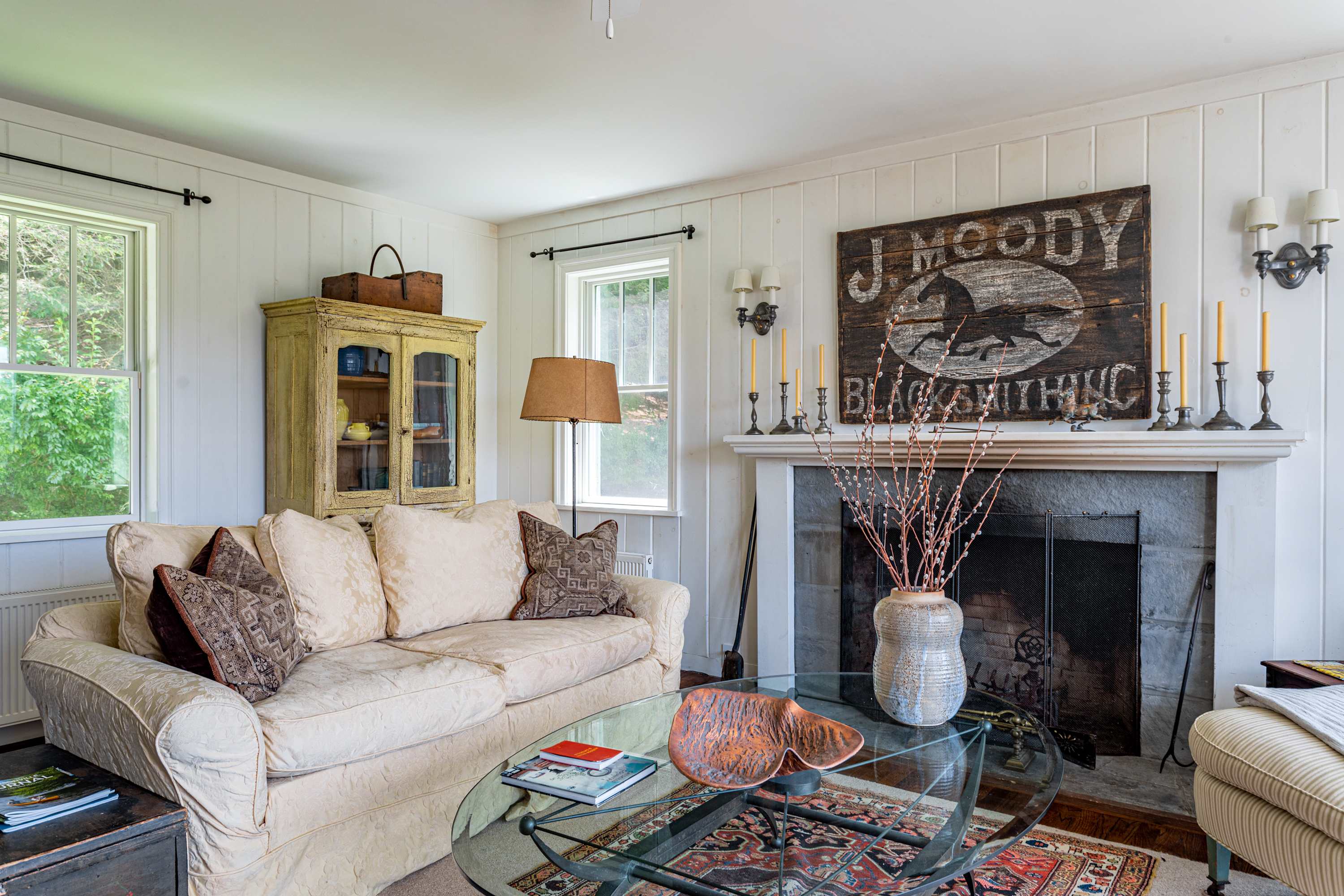 A warm living room with a stone fireplace, white paneled walls, a cream-colored sofa, and a glass coffee table, adorned with a large "J. Moody Blacksmith" sign above the mantel.
