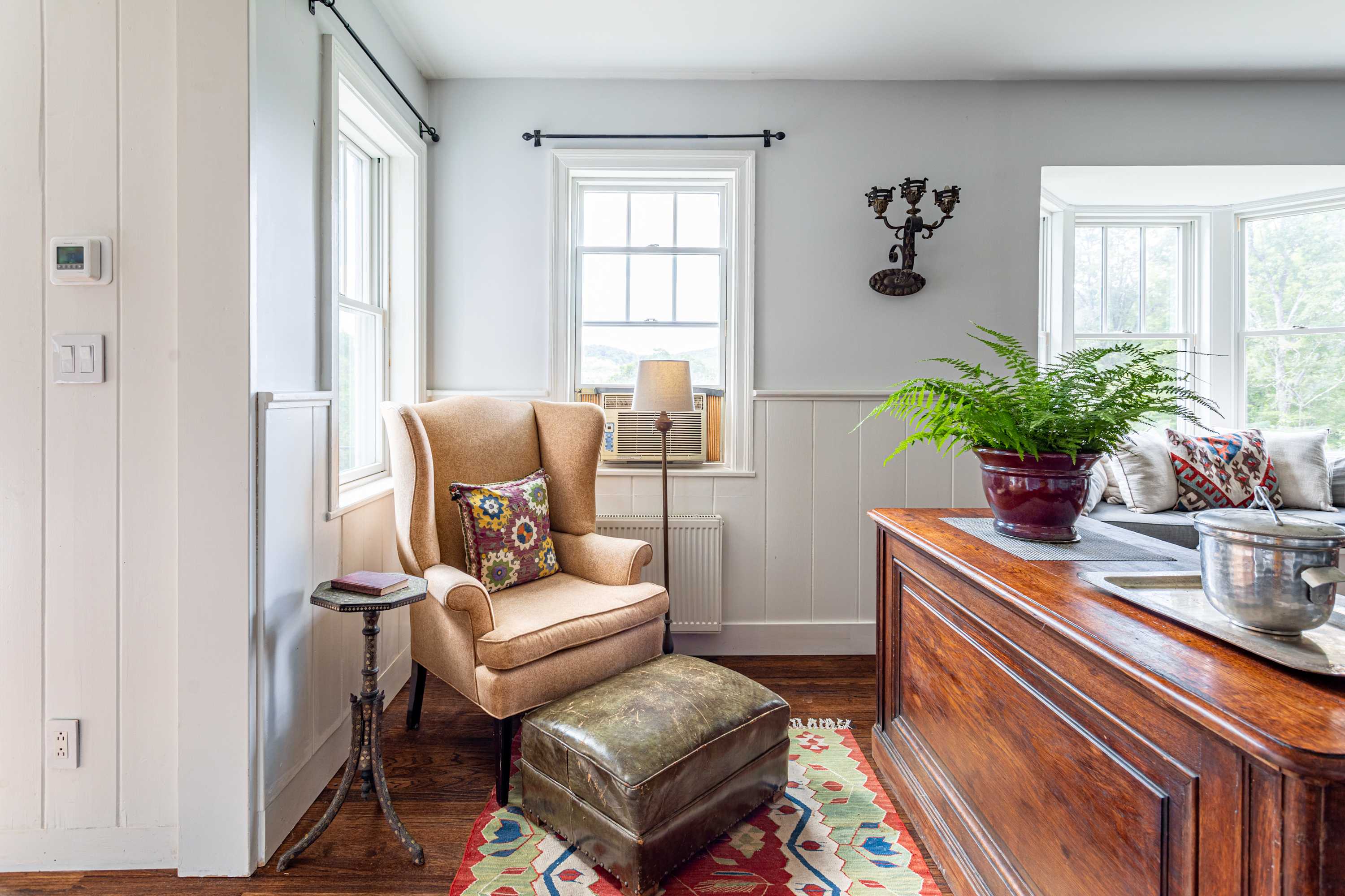 A cozy corner of the living space with a comfortable armchair and ottoman, a small side table, and a window overlooking greenery, with a colorful rug on dark hardwood floors.