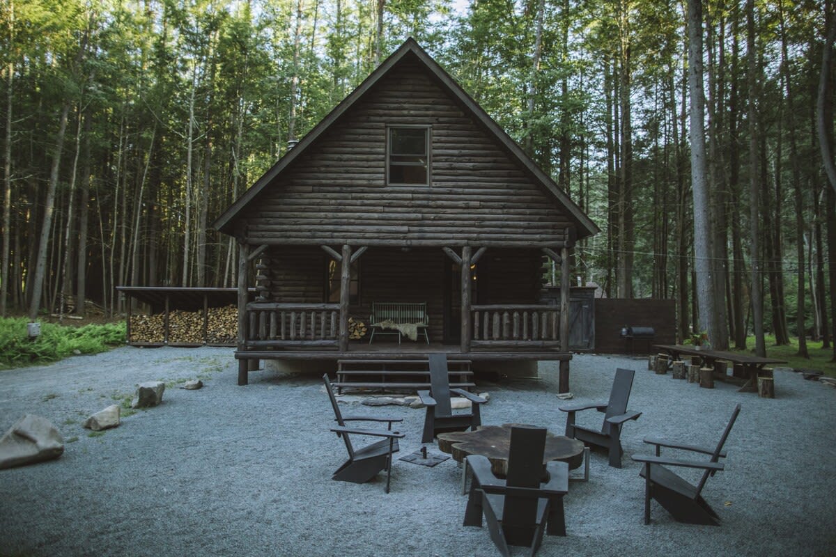 Front view of a dark log cabin with a gabled roof and a covered porch, surrounded by a dense forest. A gravel area with black Adirondack chairs and a fire pit table is in front of the cabin.