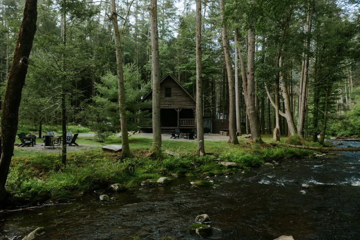 Scenic view of the log cabin nestled in a dense forest, with a river flowing in the foreground. Outdoor seating and dining areas are visible on the gravel ground near the cabin.